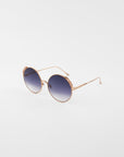 Round sunglasses with gold-colored frames and blue gradient lenses. Handmade gold-plated detailing adorns the top of the lenses. The ultra-lightweight shatter resistant lenses are 100% UVA & UVB-protected, ensuring both style and safety. The Lindy by For Art's Sake® is positioned against a plain white background.