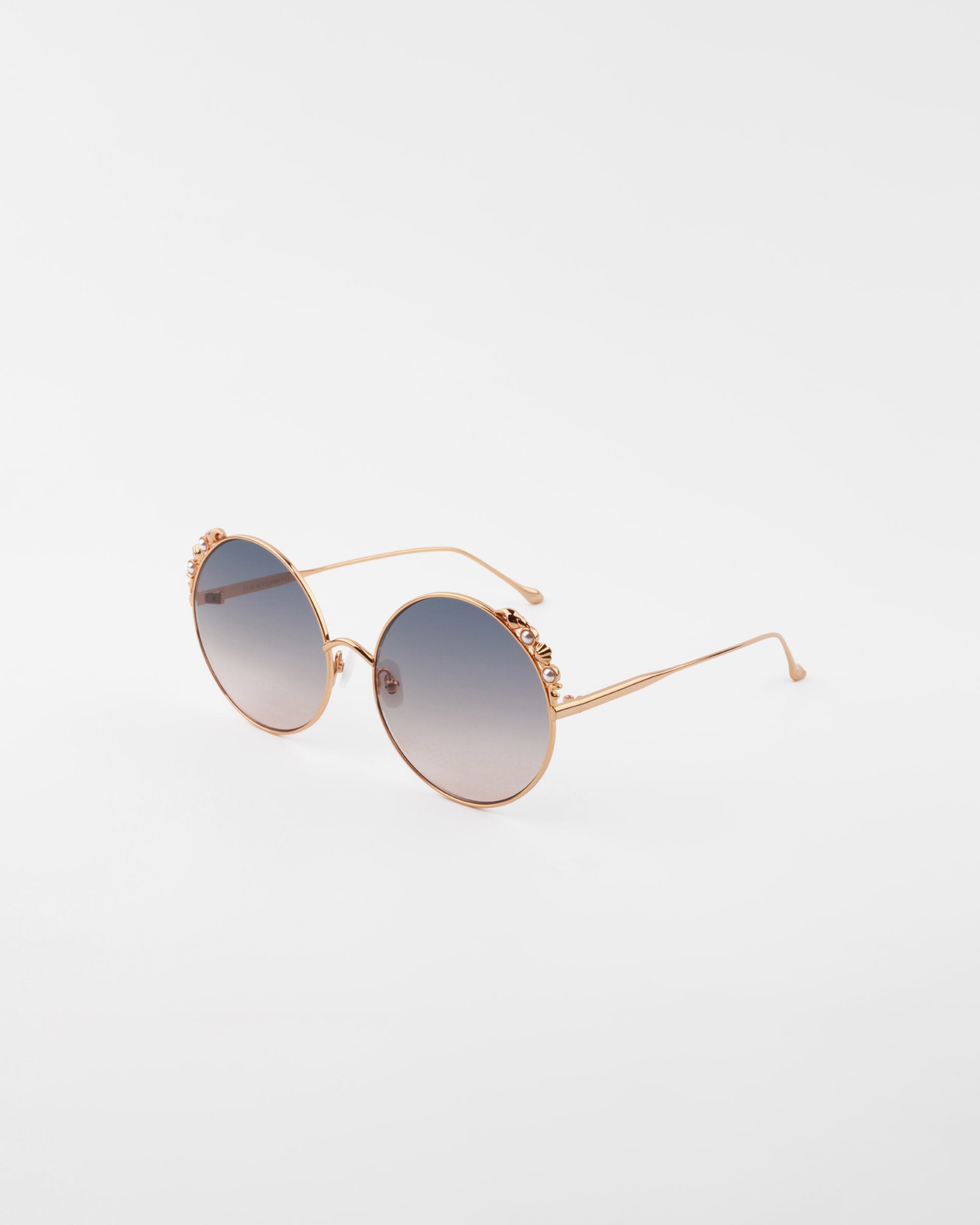 A pair of handmade gold-plated Lindy sunglasses by For Art&#39;s Sake® with round frames and blue gradient lenses are placed on a plain white background. The ultra-lightweight, shatter-resistant lenses offer 100% UVA &amp; UVB protection, while the frames feature a decorative floral design on the upper edges.