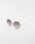 A pair of handmade gold-plated Lindy sunglasses by For Art's Sake® with round frames and blue gradient lenses are placed on a plain white background. The ultra-lightweight, shatter-resistant lenses offer 100% UVA & UVB protection, while the frames feature a decorative floral design on the upper edges.