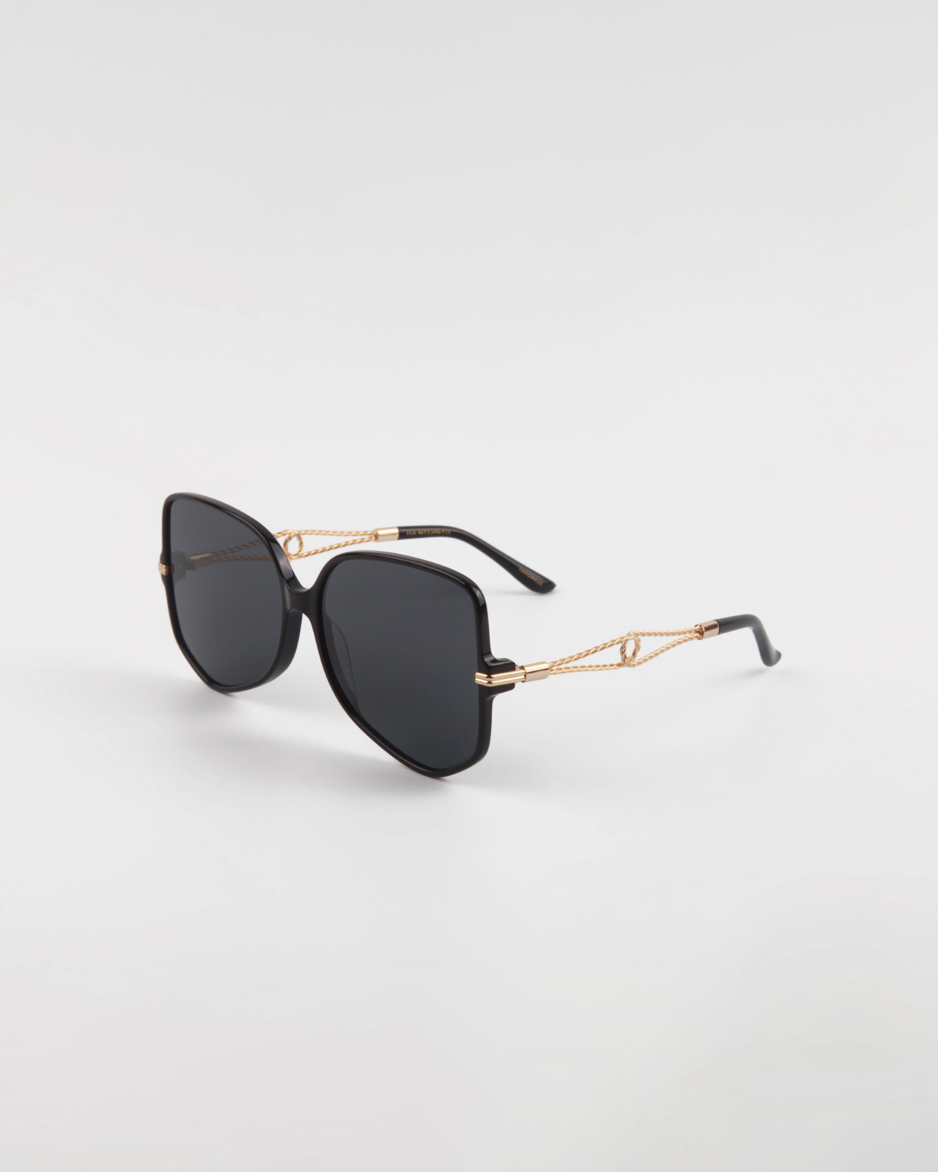 A pair of black Voyager sunglasses from For Art's Sake® with large, square lenses and gold-tone intricate detailing on the temples are placed against a plain white background. The arms of the Voyager sunglasses, crafted from handmade acetate eyewear, have a slight curve towards the ends.