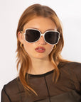 A person with long, light brown hair is wearing large, square-shaped white Voyager sunglasses from For Art's Sake® made from plant-based acetate with dark lenses that offer UV protection. They are also adorned with gold hoop earrings and a black, semi-sheer top. The background is white.