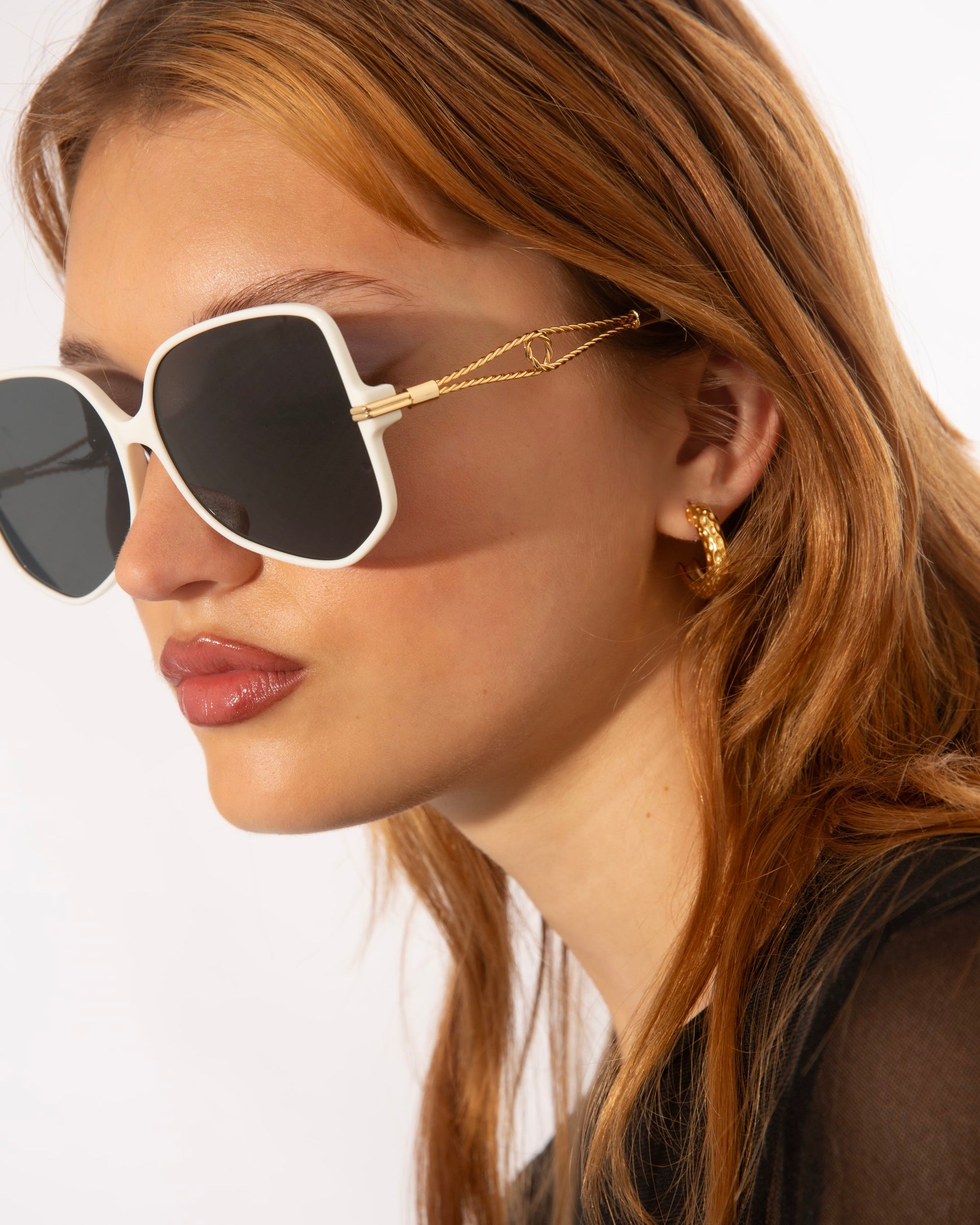 A person with long, auburn hair wearing square, oversized For Art's Sake® Voyager sunglasses with black lenses and 18-karat gold-plated chains is shown in a close-up profile view. They have light skin and are also wearing gold hoop earrings. The background is plain and white.