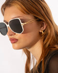 A person with long, auburn hair wearing square, oversized For Art's Sake® Voyager sunglasses with black lenses and 18-karat gold-plated chains is shown in a close-up profile view. They have light skin and are also wearing gold hoop earrings. The background is plain and white.