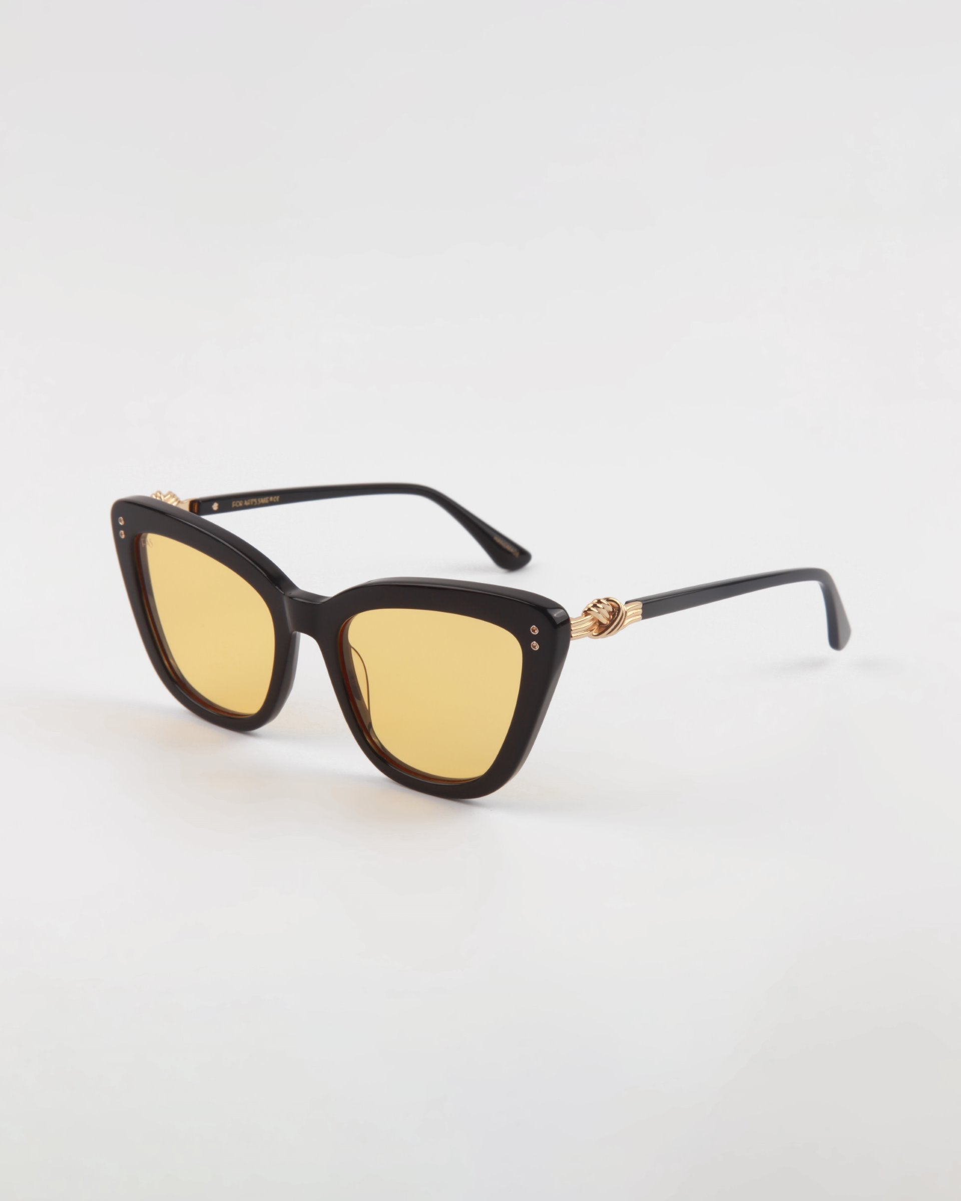 Ice Cream by For Art&#39;s Sake® with black handmade acetate cat-eye frames and yellow-tinted lenses. The frames feature gold decorative elements on the hinges, providing both style and essential UVA &amp; UVB protection. These chic glasses are elegantly displayed on a minimalist white background.
