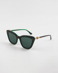 A pair of stylish, dark green cat-eye frames crafted from handmade acetate with dark tinted lenses. The arms feature a gold twist detail near the hinges, adding an elegant touch. These Ice Cream sunglasses by For Art's Sake® provide complete UVA & UVB protection, ensuring both style and safety. The background is plain white.