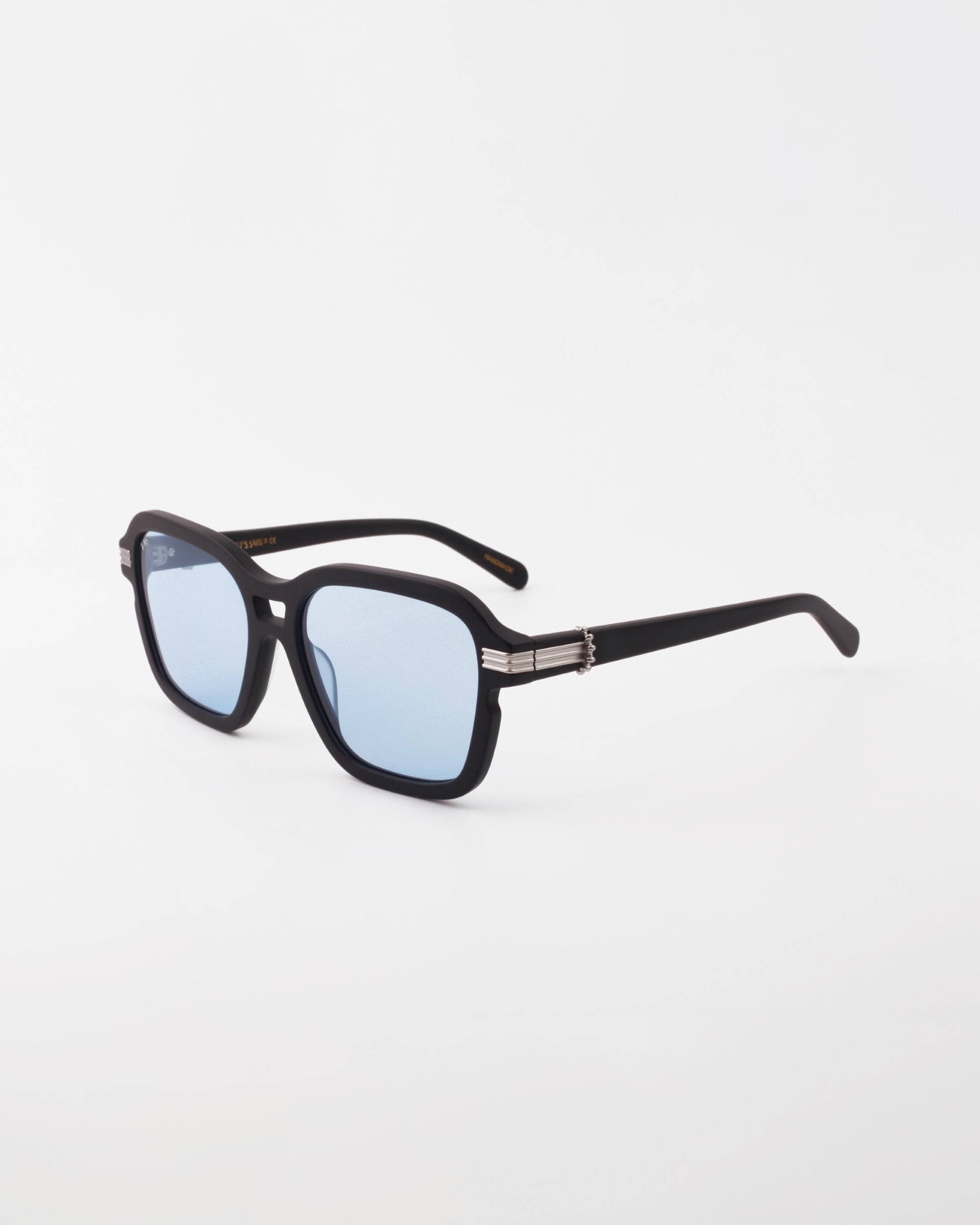 A pair of black-framed Shadyside sunglasses from For Art&#39;s Sake® with light blue, shatter-resistant nylon lenses. The hand-polished acetate arms feature a distinctive metallic decorative element near the hinges. The Shadyside sunglasses are placed against a plain white background.