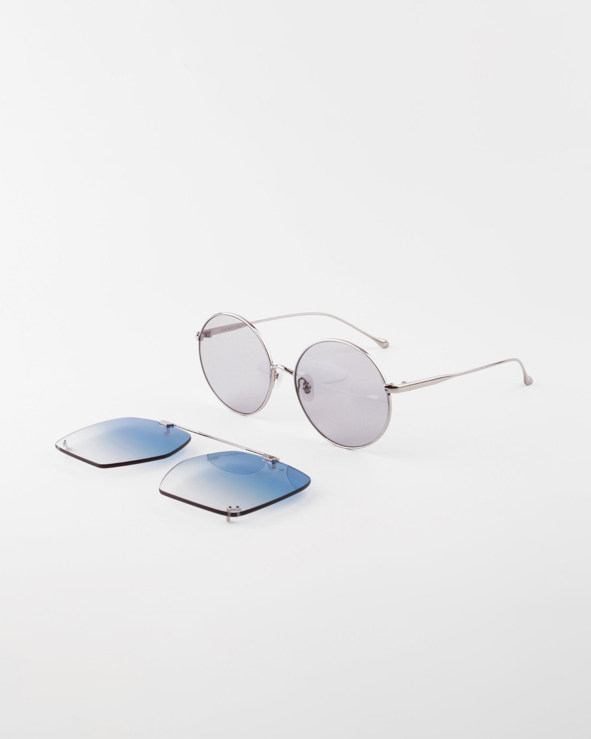 A pair of round, silver-framed eyeglasses with detachable blue-tinted sun lenses on a white background. The For Art's Sake® Last Summer detachable aviator frame offers reversible functionality, with the blue-tinted lenses positioned in front of the glasses for UVA & UVB protection.