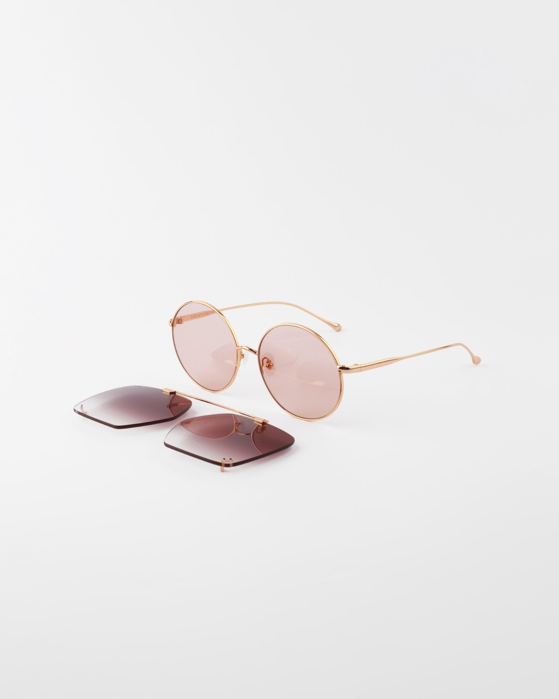A pair of round-framed, pink-tinted Last Summer sunglasses by For Art&#39;s Sake® with an 18-karat gold-plated wire frame is placed on a white surface. Adjacent to the Last Summer sunglasses by For Art&#39;s Sake® are a UVA &amp; UVB-protected pair of square, dark-tinted lenses.