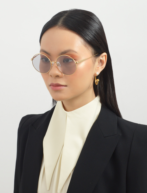 A person with long dark hair wearing round, UVA &amp; UVB-protected For Art&#39;s Sake® Last Summer eyeglasses, gold hoop earrings, and a black blazer over a white blouse with a wide collar. The background is plain white.
