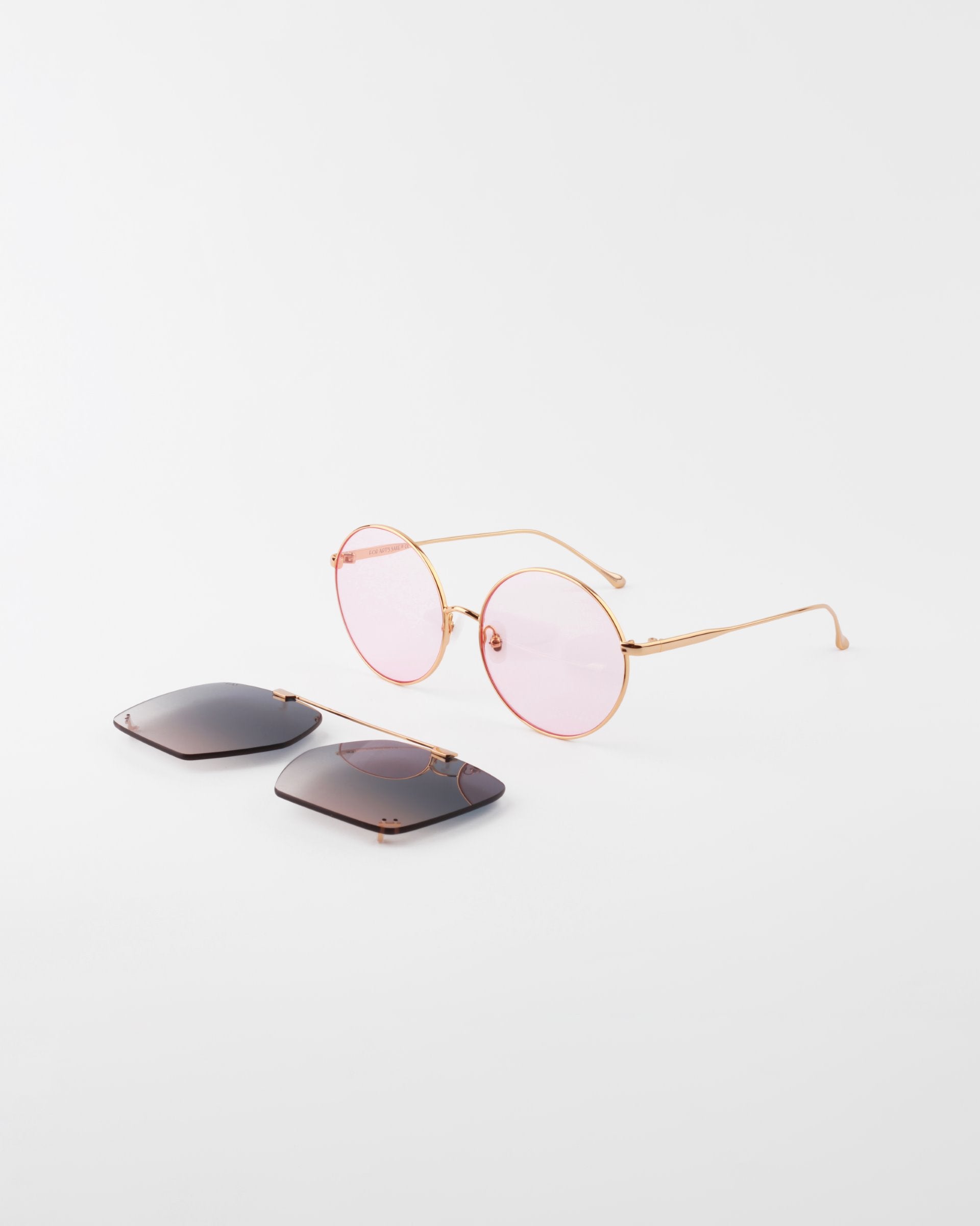 A pair of round, 18-karat gold-plated eyeglasses with light pink lenses, called &quot;Last Summer&quot; by For Art&#39;s Sake®, are displayed on a white surface. Beside the glasses, there are two rectangular dark gray clip-on lenses unattached, offering additional UVA &amp; UVB protection.