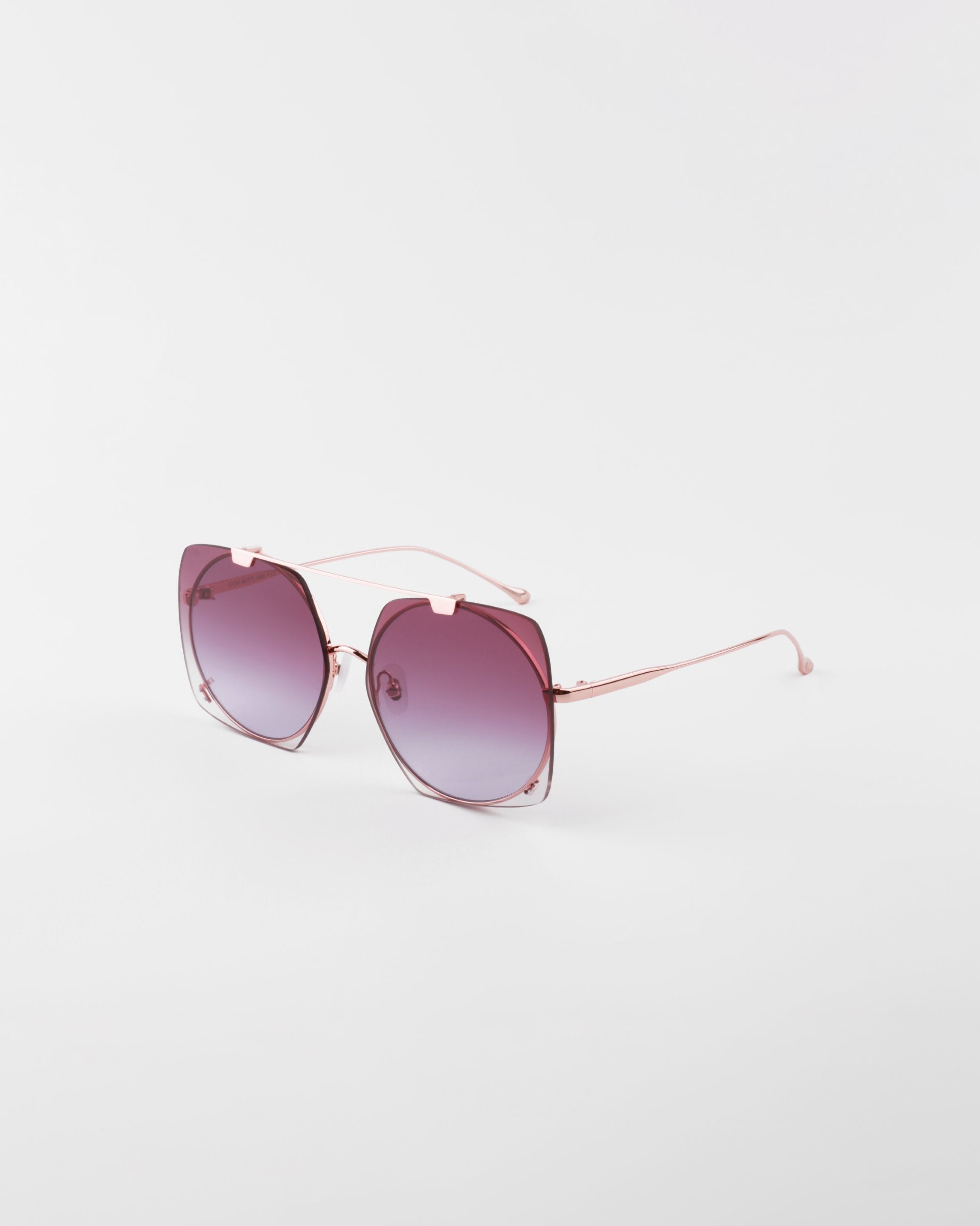 A pair of stylish Last Summer sunglasses by For Art&#39;s Sake® with rose-tinted lenses and thin, metallic pink frames, UVA &amp; UVB-protected. The sunglasses are positioned on a white surface, showcasing a gradient on the lenses from dark at the top to lighter at the bottom.