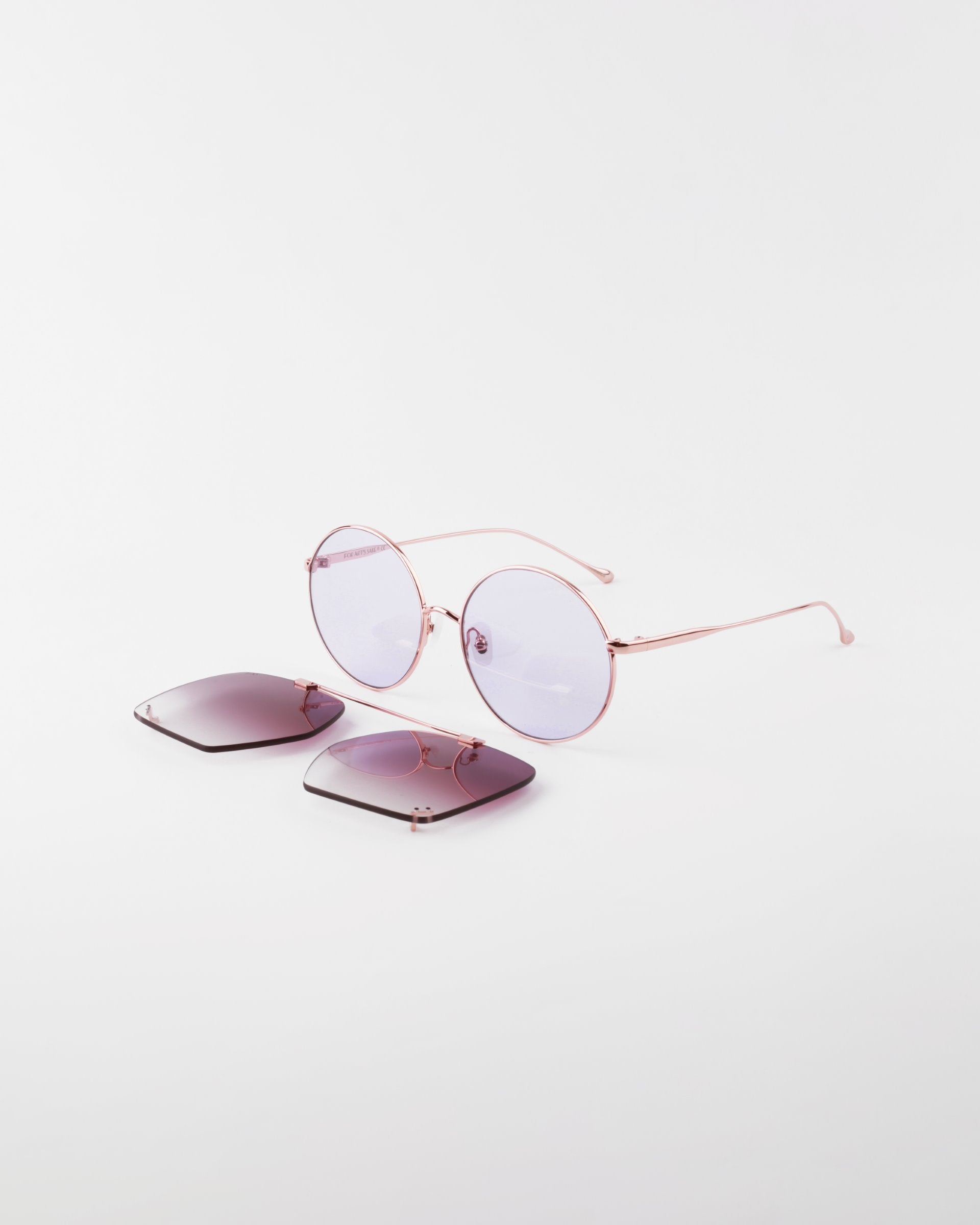 A pair of 18-karat gold-plated, round-frame Last Summer sunglasses by For Art&#39;s Sake® with removable purple lenses sit on a white surface. One set of lens is detached and laid flat nearby, showcasing the sunglasses&#39; interchangeable lens feature and UVA &amp; UVB-protected design.