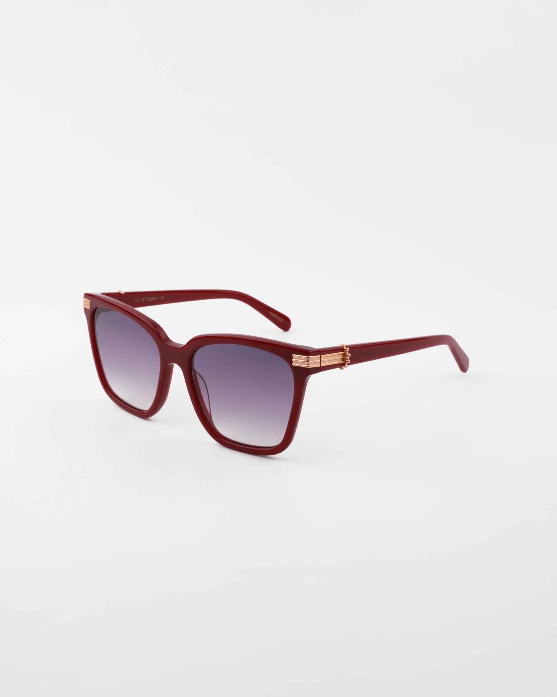 A pair of stylish, handmade acetate sunglasses featuring dark maroon frames and gradient purple, UV-protected lenses. The temples boast an 18-karat gold-plated decorative accent near the hinges. The Gaia sunglasses by For Art&#39;s Sake® are positioned on a white background.