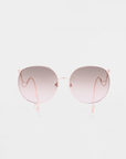 A pair of For Art's Sake® Birthday Cake sunglasses with handmade gold-plated, rose gold frames and gradient lenses that transition from light pink at the bottom to darker pink at the top. The UVA & UVB-protected, shatter-resistant lenses offer clear sight while delicate, curved arms and clear nose pads add elegance. The background is plain white.