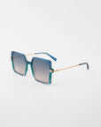 Square-shaped handmade acetate Moxie sunglasses by For Art's Sake® with gradient green and blue frames and 18-karat gold-plated temples on a white background. The lenses are tinted, transitioning from light to dark towards the top, offering UVA & UVB protection. The temple tips are dark blue.