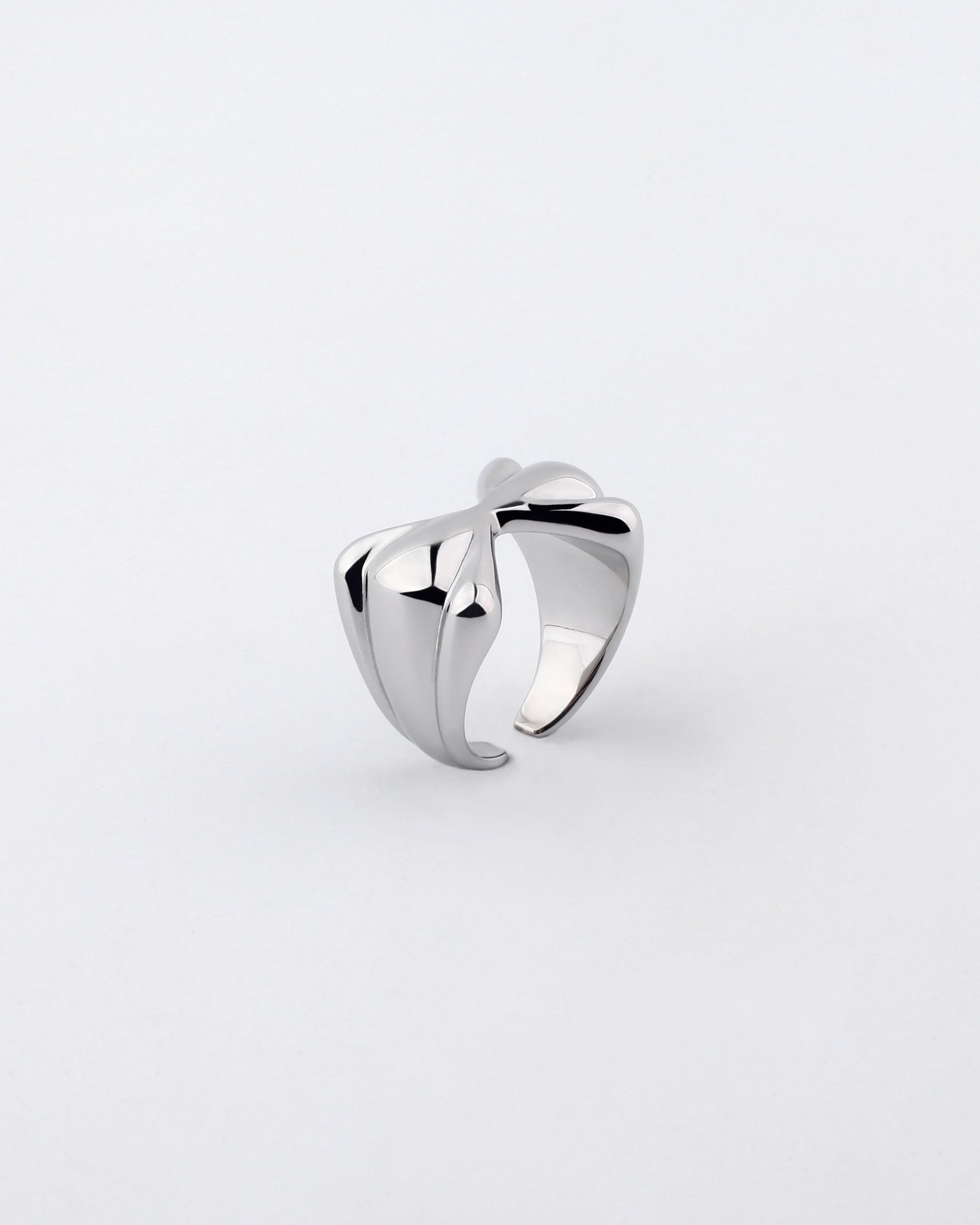 The Sydney Ring by For Art's Sake® features a polished, silver-plated finish and showcases a contemporary bow-shaped design with smooth, curved lines that intertwine to create an open and elegant form. Its surface reflects light, highlighting the sleek and modern aesthetic against a plain white background.