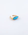 The Enza Ring from For Art's Sake® features a gold-plated stainless steel open band design, adorned with an oval-shaped turquoise gemstone set on top. The piece is displayed against a plain white background, accentuating its polished enamel finish.