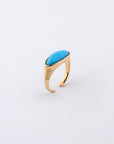 The Enza Ring by For Art's Sake®, made of gold-plated stainless steel, features an open band design with a striking elongated oval turquoise stone at its center. The band is adorned with delicate horizontal etched lines, adding texture and creating a modern yet elegant look against a plain white background.