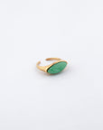 The Enza Ring from For Art's Sake® features a prominent oval-shaped green stone set in a gold-plated stainless steel band. The band gracefully widens around the stone, boasting a sleek, modern design with a polished enamel finish. It is displayed against a plain, light gray background.