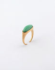 The Enza Ring by For Art's Sake® is a gold-plated stainless steel ring adorned with an elongated oval green gemstone on top, shown against a plain white background. It showcases a sleek, modern design with grooved detailing on the band and a polished enamel finish for added elegance.