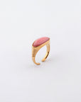 The Enza ring by For Art's Sake® is a gold open-ended piece featuring an elongated oval coral-colored stone set horizontally at the top. Crafted from gold-plated stainless steel, it boasts a minimalist design with a polished finish and subtle ridged detailing on the band near the stone, set against a white background.