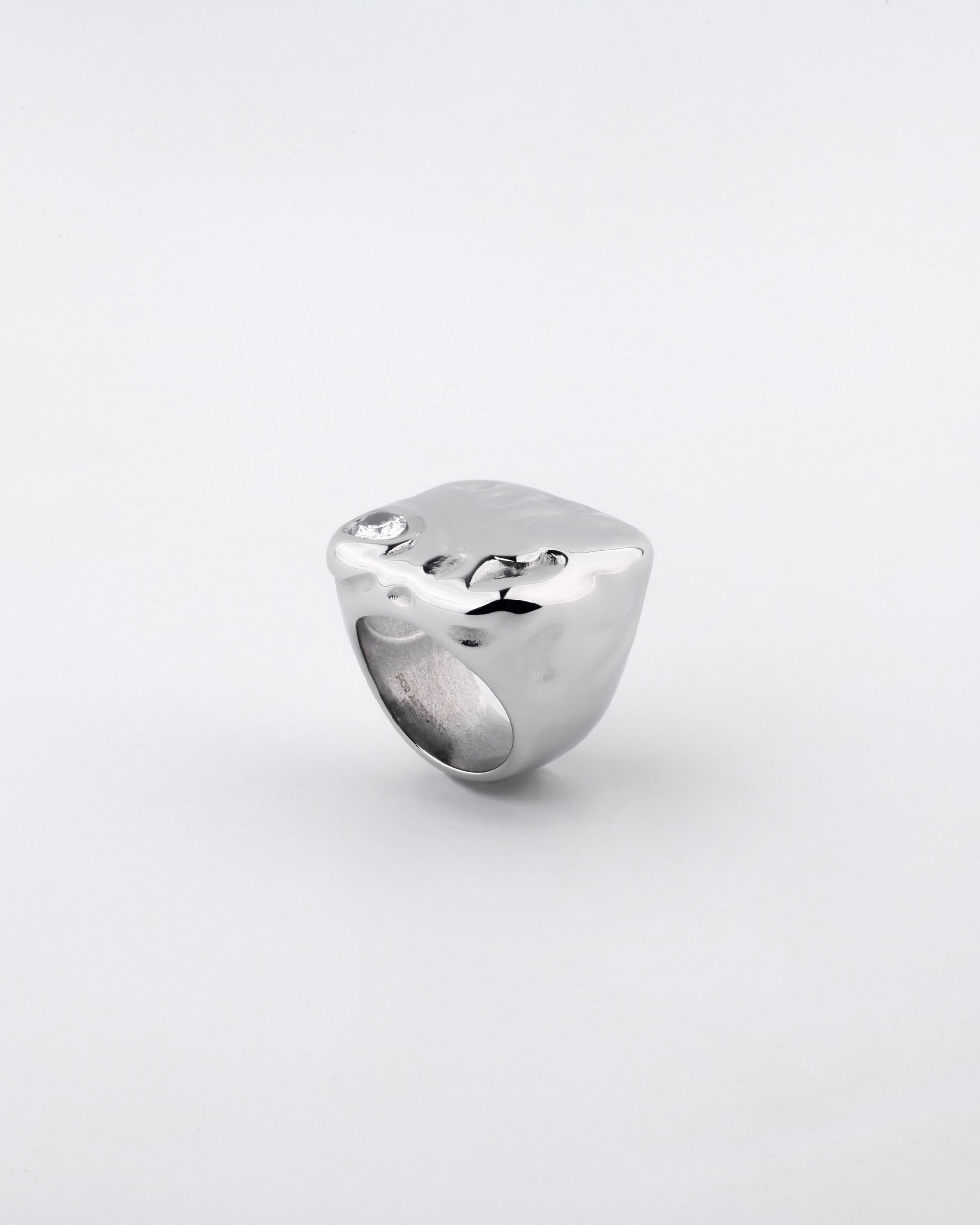 Introducing the Chiara Ring by For Art's Sake®: a polished, silver-plated masterpiece. This ring boasts a thick, square-shaped band with smooth, rounded edges and features an eye-catching abstract wavy surface. A small, sparkling gemstone embedded near one corner adds a touch of elegance against its plain white backdrop.