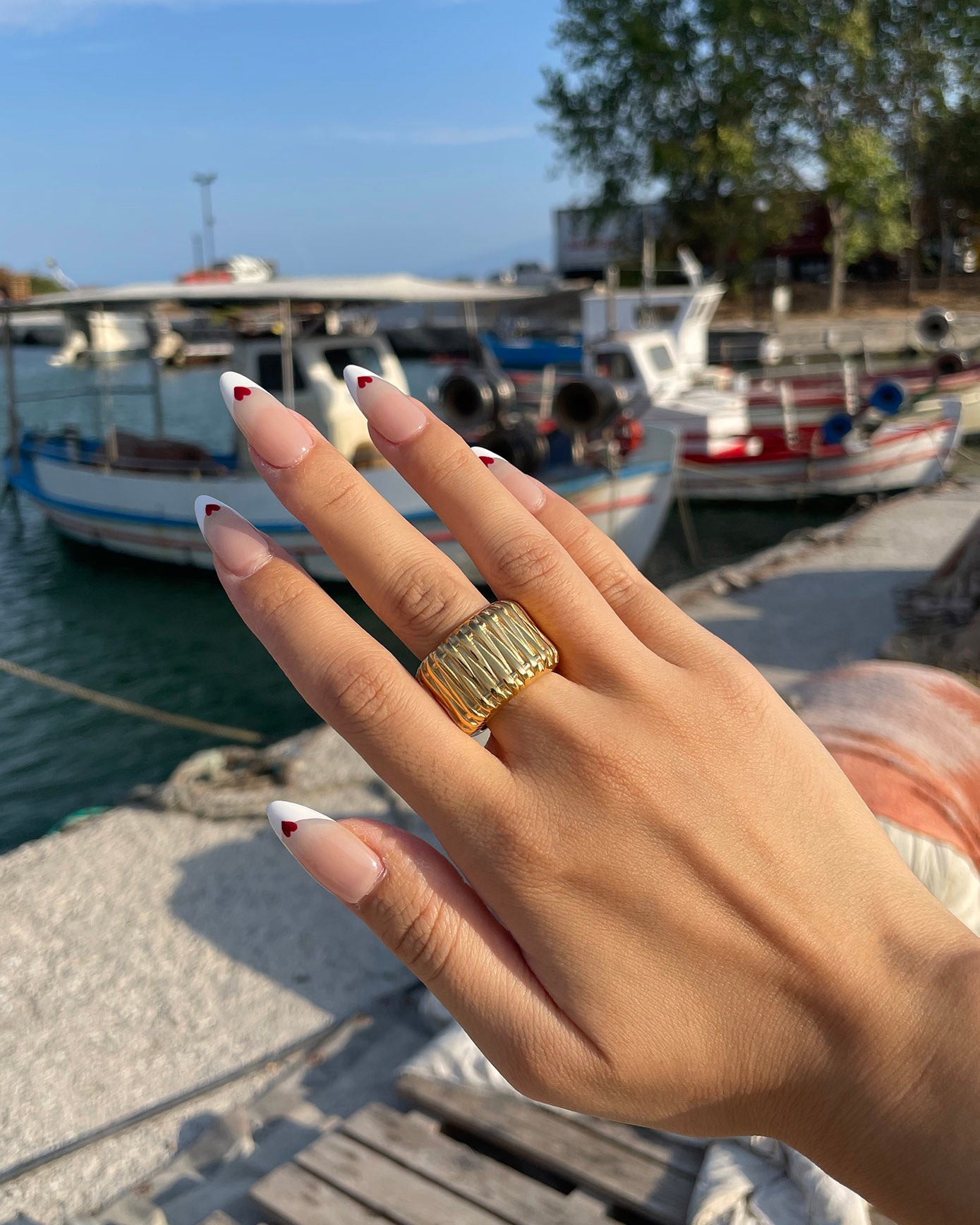 A hand adorned with the For Art's Sake® Woven Ring, showcasing artistic craftsmanship and manicured nails featuring red-tipped nail art, is held up against a backdrop of boats docked at a marina. The water is calm and the sky is clear, with trees visible in the background.