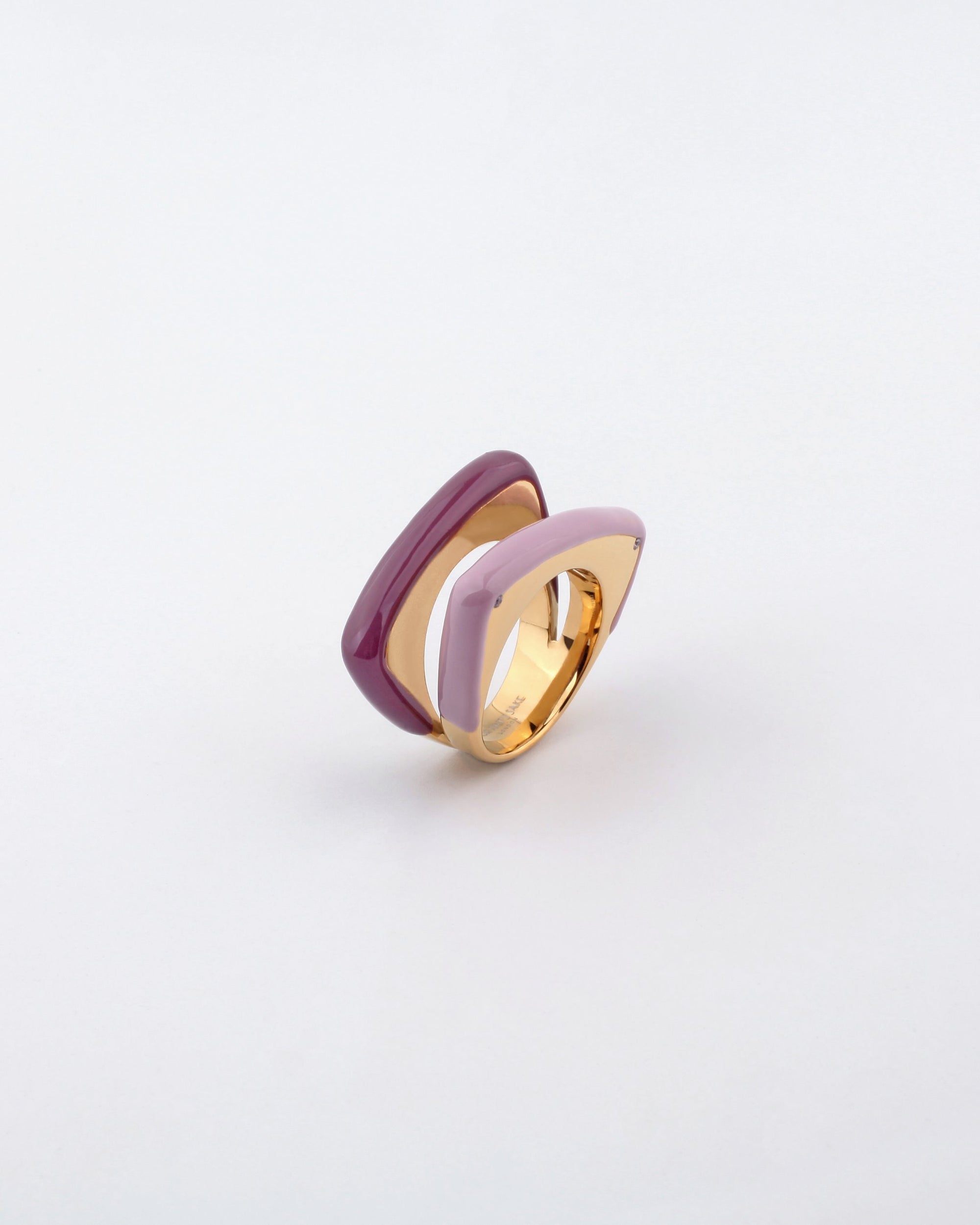 The Delta Ring by For Art&#39;s Sake® showcases a modern layered design featuring two geometric shapes coated in enamel. One shape boasts a deep, glossy maroon hue while the other displays a soft, pastel lavender, creating an eye-catching contrast. The contemporary aesthetic of this gold ring is beautifully highlighted against a plain white background.