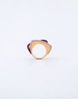 The Delta Ring by For Art's Sake® is a uniquely designed piece featuring a large circular hole in the center. Its abstract shape combines shades of purple and gold with a matte finish. The layered design appears modern and artistic, creating a striking visual contrast against the plain white background.