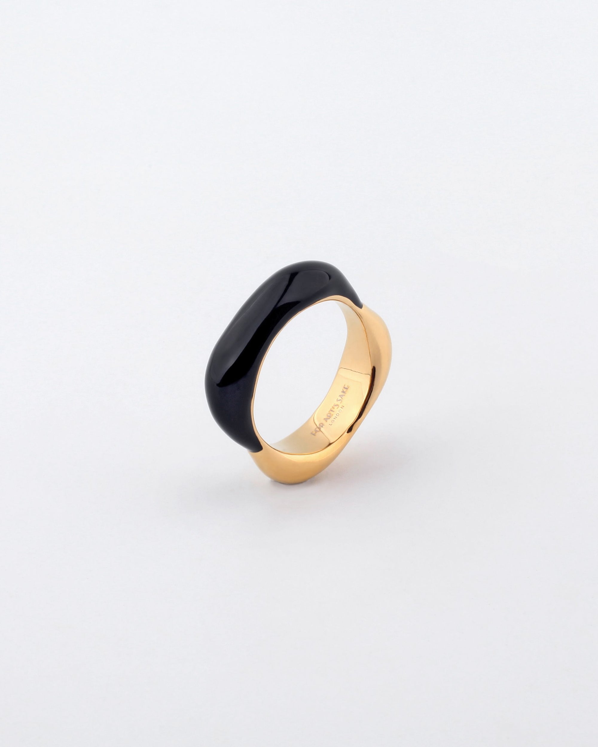 A Raya Ring from For Art's Sake® with a large, smooth black oval stone embedded in it is displayed on a white background. The inner band features an engraved inscription. This gold-toned metal ring has a modern, elegant design.