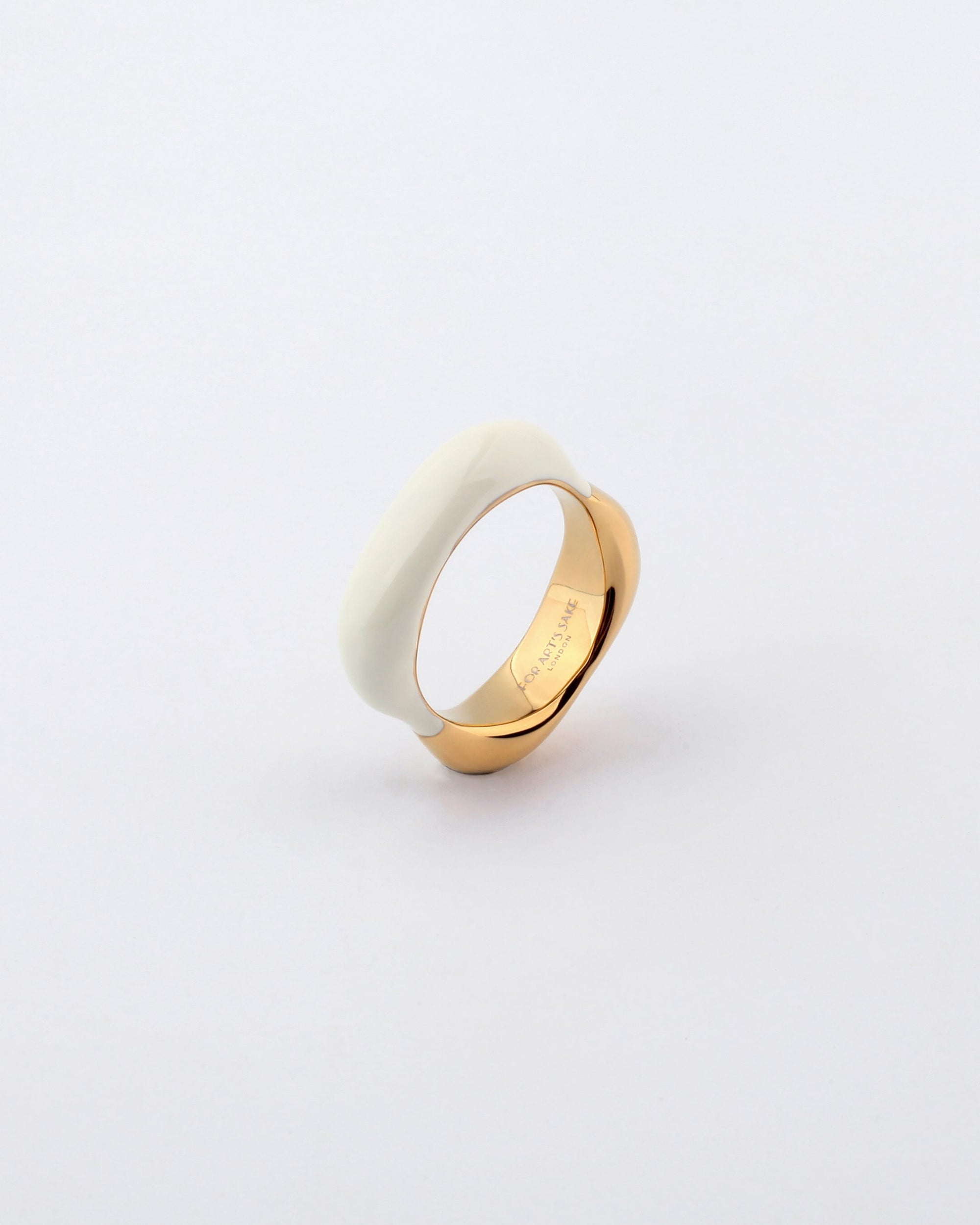 The Raya Ring by For Art's Sake® is a piece of contemporary jewellery, uniquely designed with a fusion of two contrasting materials: a smooth white ceramic or resin band seamlessly intertwined with a polished gold-colored metal band, set against a plain light grey background.