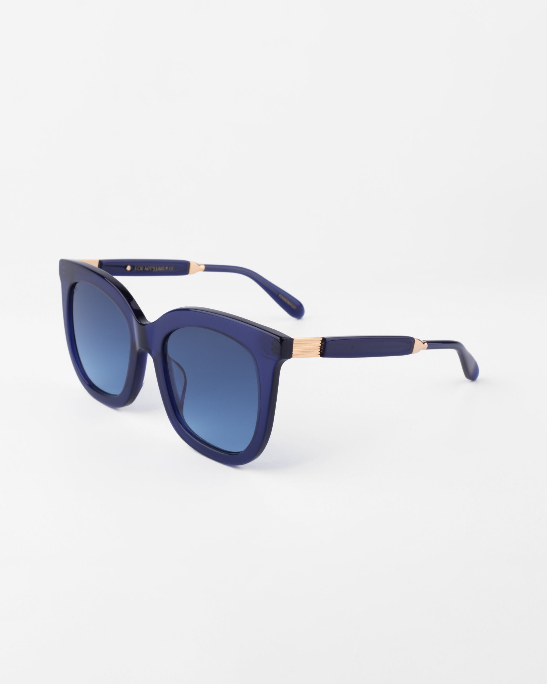 A pair of Riverside sunglasses by For Art's Sake® with square, shatter-resistant, dark blue lenses and a handmade acetate frame. The temples also feature gold-plated detailing and are similarly dark blue, adding an elegant touch to the design. The Riverside sunglasses are set against a plain white background.
