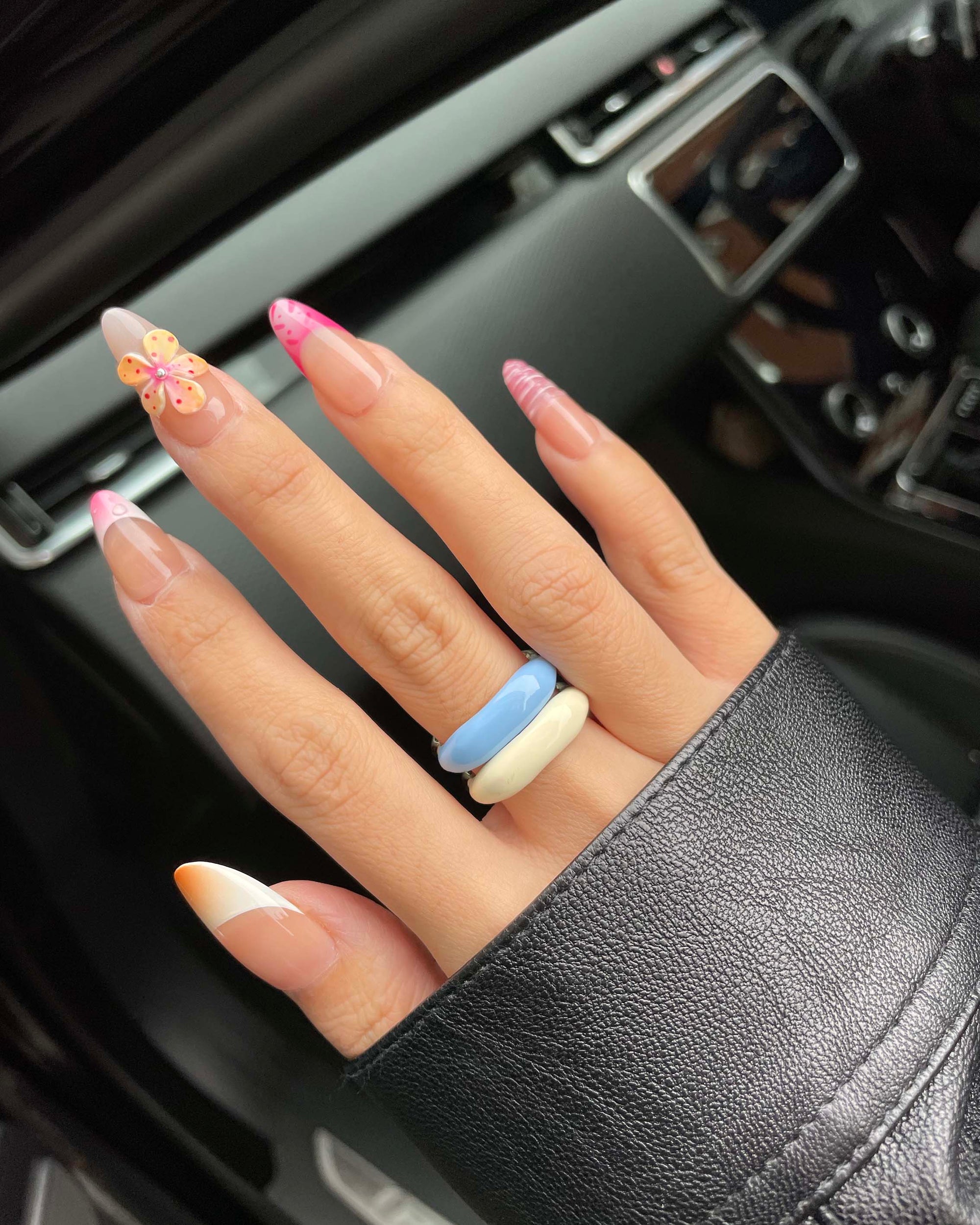 A hand with vibrant nail art showcasing a butterfly design on the ring finger is elegantly adorned with a minimalist Raya Ring by For Art&#39;s Sake®. The individual wears a black leather jacket accompanied by two chunky rings in shades of blue and cream. The backdrop displays the sleek interior of a car.