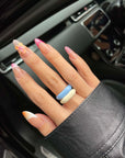 A hand adorned with vibrant nail art, showcasing pink tips, a butterfly motif, and striped patterns, is positioned on a car steering wheel. The individual wears chunky Raya Rings by For Art's Sake® in shades of blue and cream over an enamel finish and a black leather sleeve.