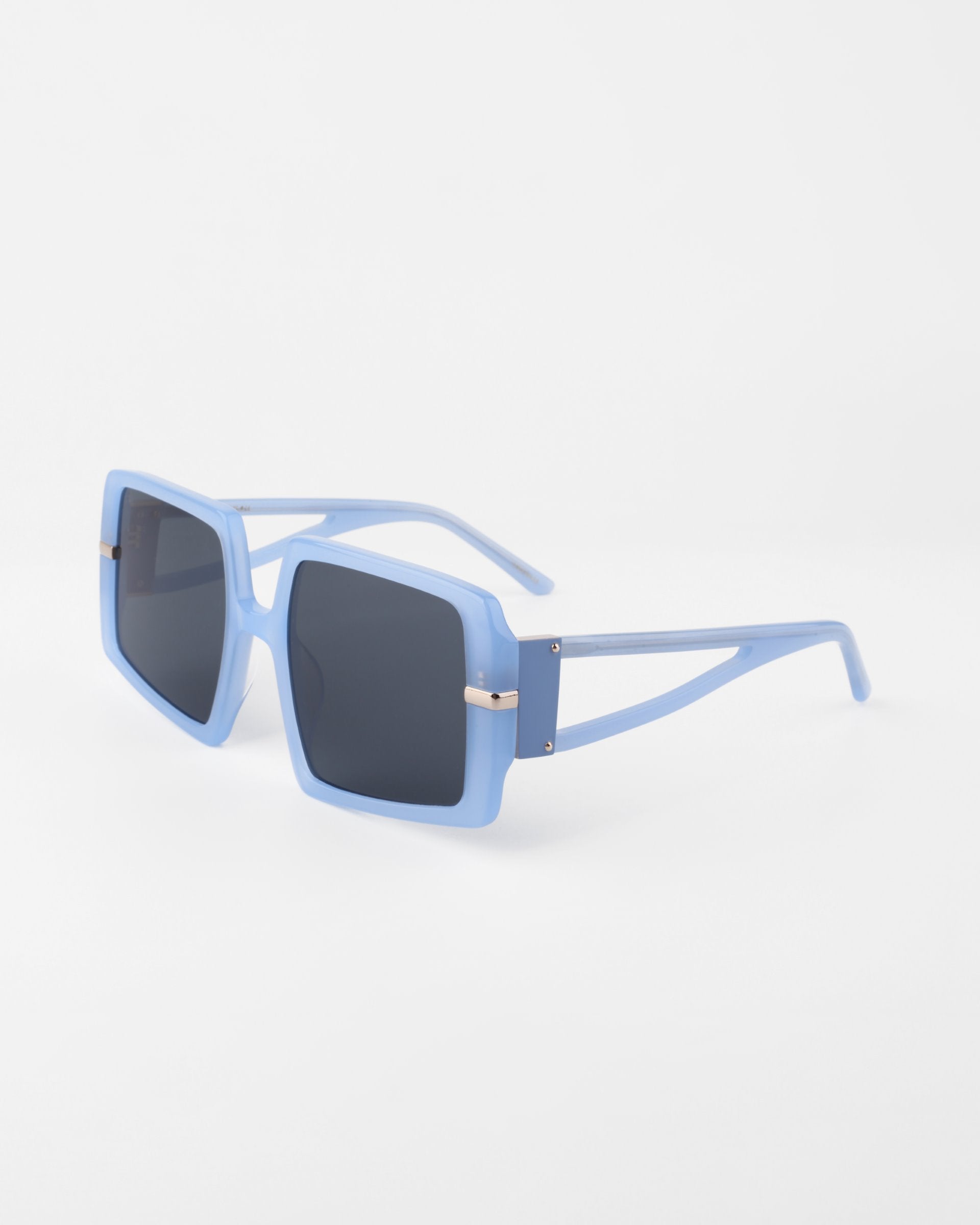 A pair of stylish, square-framed For Art&#39;s Sake® Saturday sunglasses with light blue rims and dark tinted shatter-resistant nylon lenses. The arms of the sunglasses also feature light blue color with a thin, cut-out design near the hinges, which include small metallic accents.