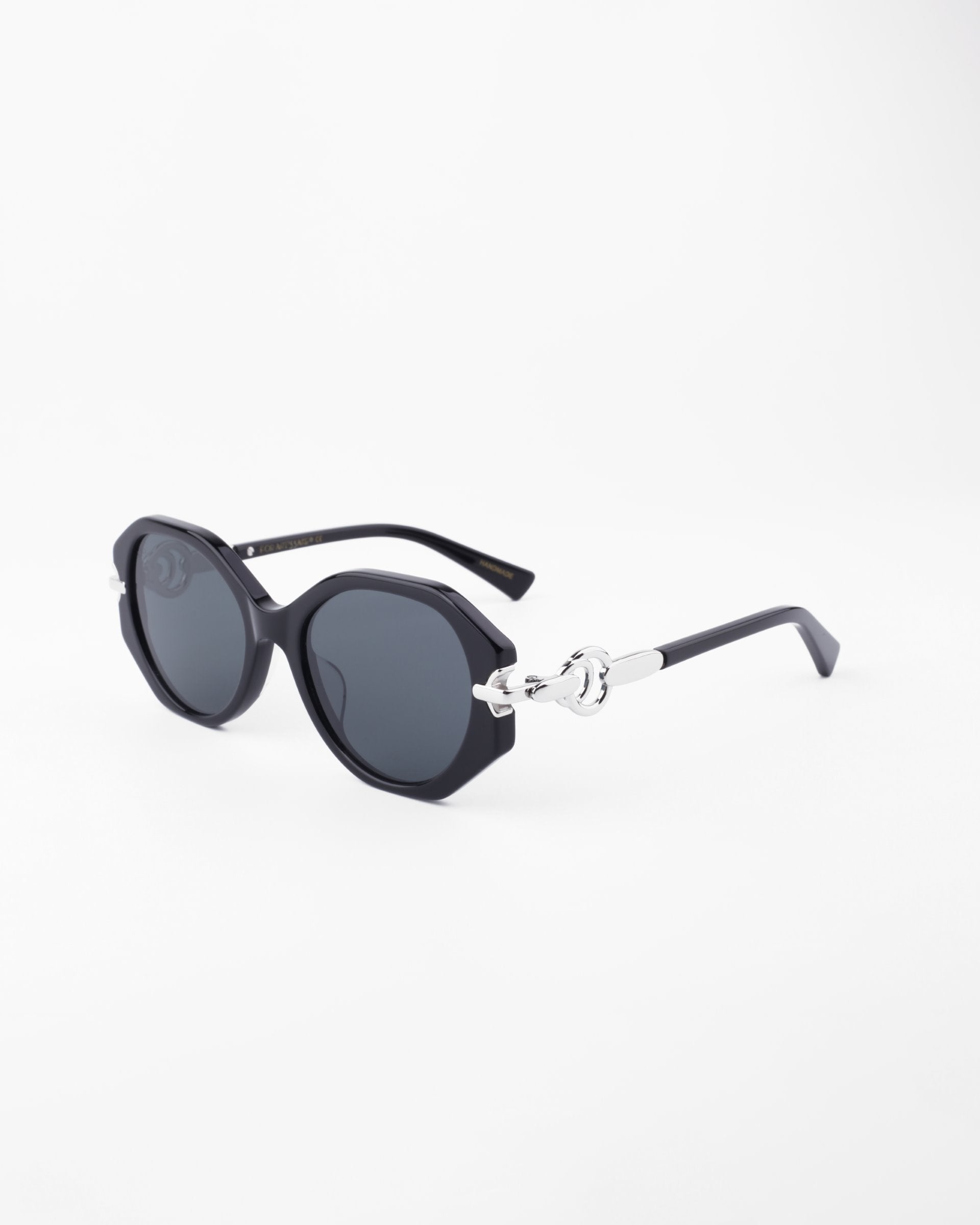 A pair of stylish black Seaside sunglasses from For Art's Sake® featuring octagonal, shatter-resistant lenses and silver accents on the hinges and arms. The design, with its modern, slightly thick rims and handmade acetate frame, exudes a fashionable and chic appearance. The background is a plain, light color.