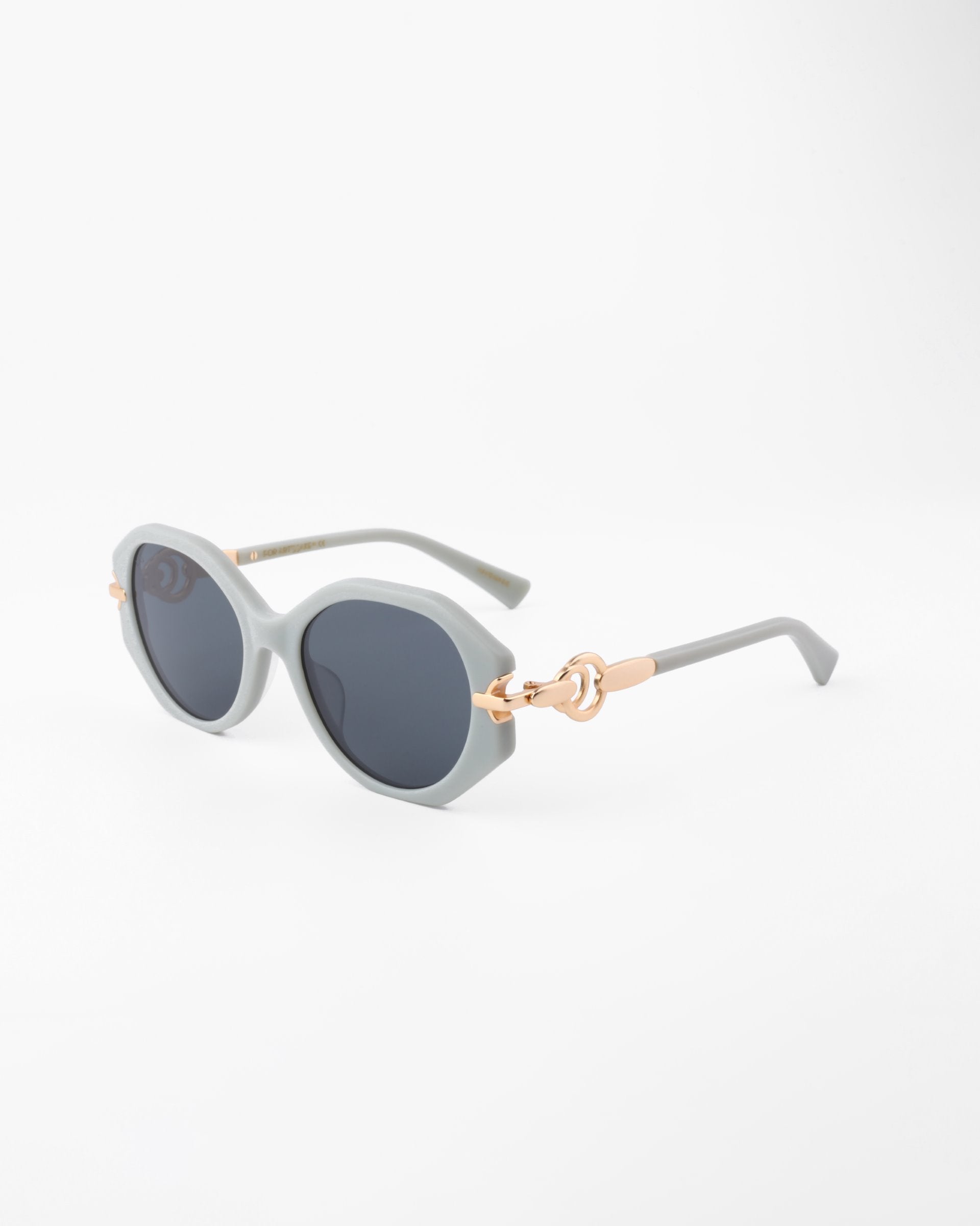 A pair of stylish Seaside sunglasses by For Art&#39;s Sake® featuring round black lenses with a light gray, semi-translucent, handmade acetate frame. The temples have gold-plated detailing near the hinges, adding a touch of elegance to the design. The background is plain white.