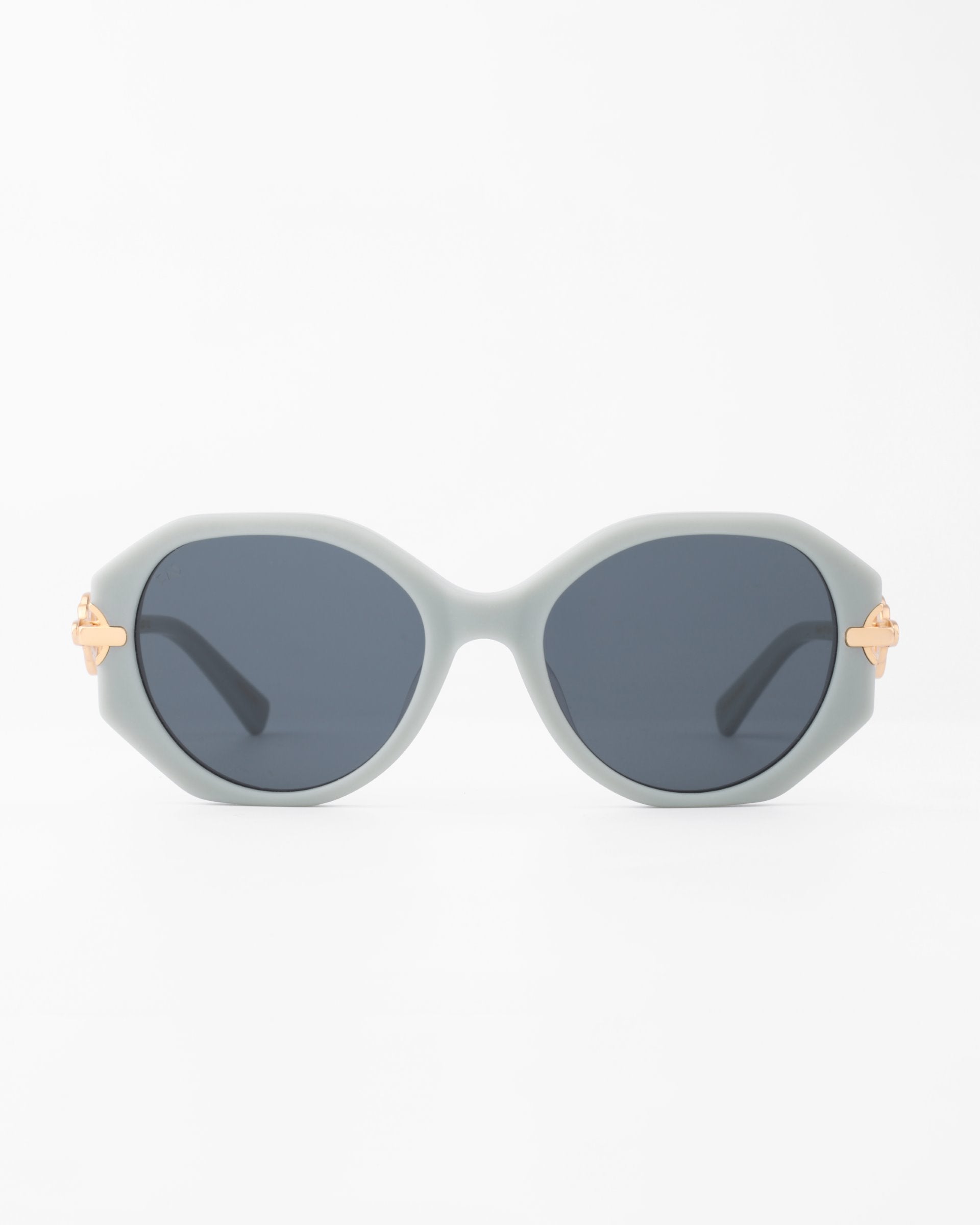 A pair of light gray oval-shaped Seaside sunglasses by For Art&#39;s Sake® with dark blue, shatter-resistant lenses. The handmade acetate frame features gold-plated temple detailing on a plain white background.