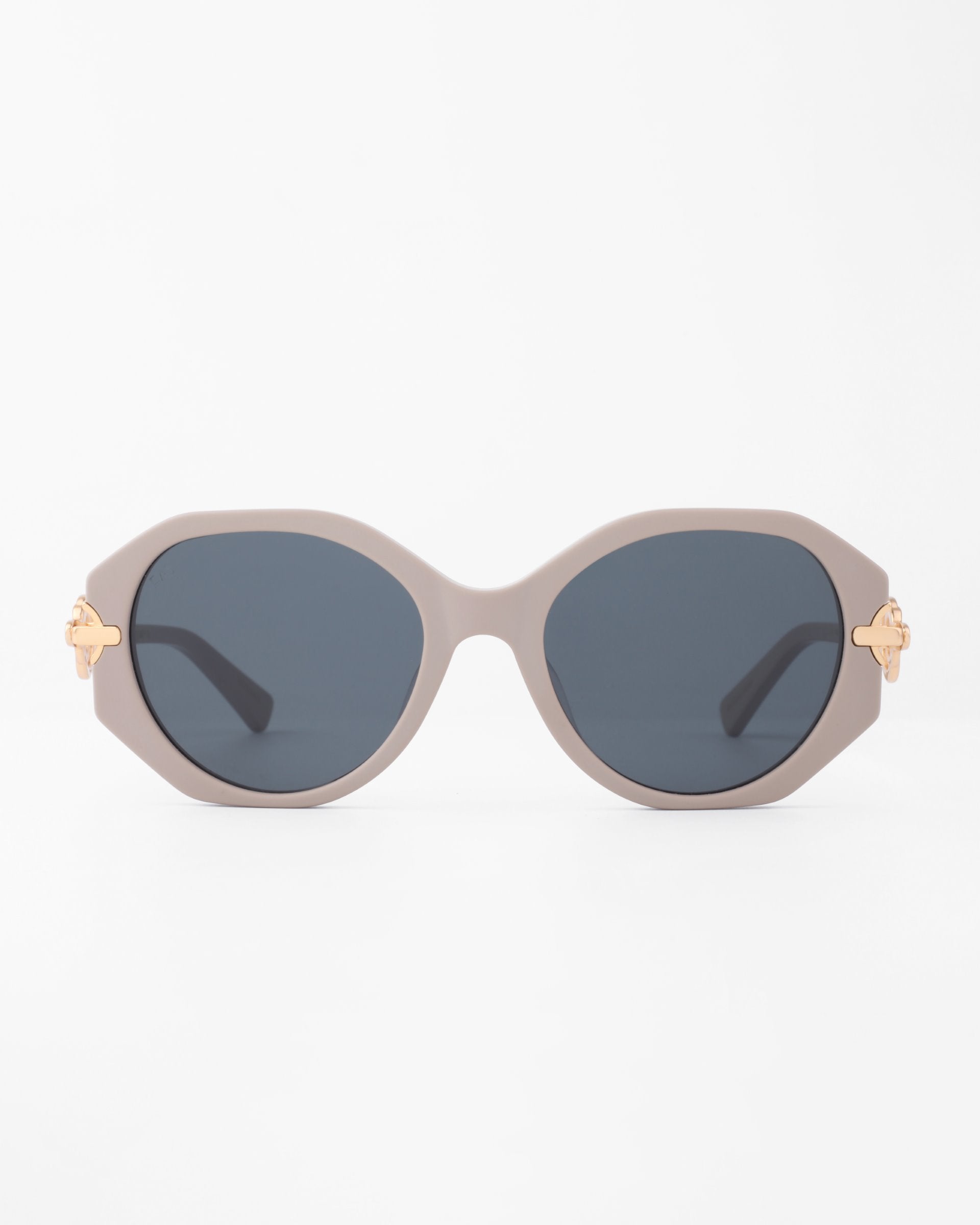A pair of stylish Seaside sunglasses by For Art&#39;s Sake® with dark oval, shatter-resistant lenses and a light beige, handmade acetate frame. The temples feature gold-plated detailing near the hinges, adding a touch of elegance to the design. The background is solid white.