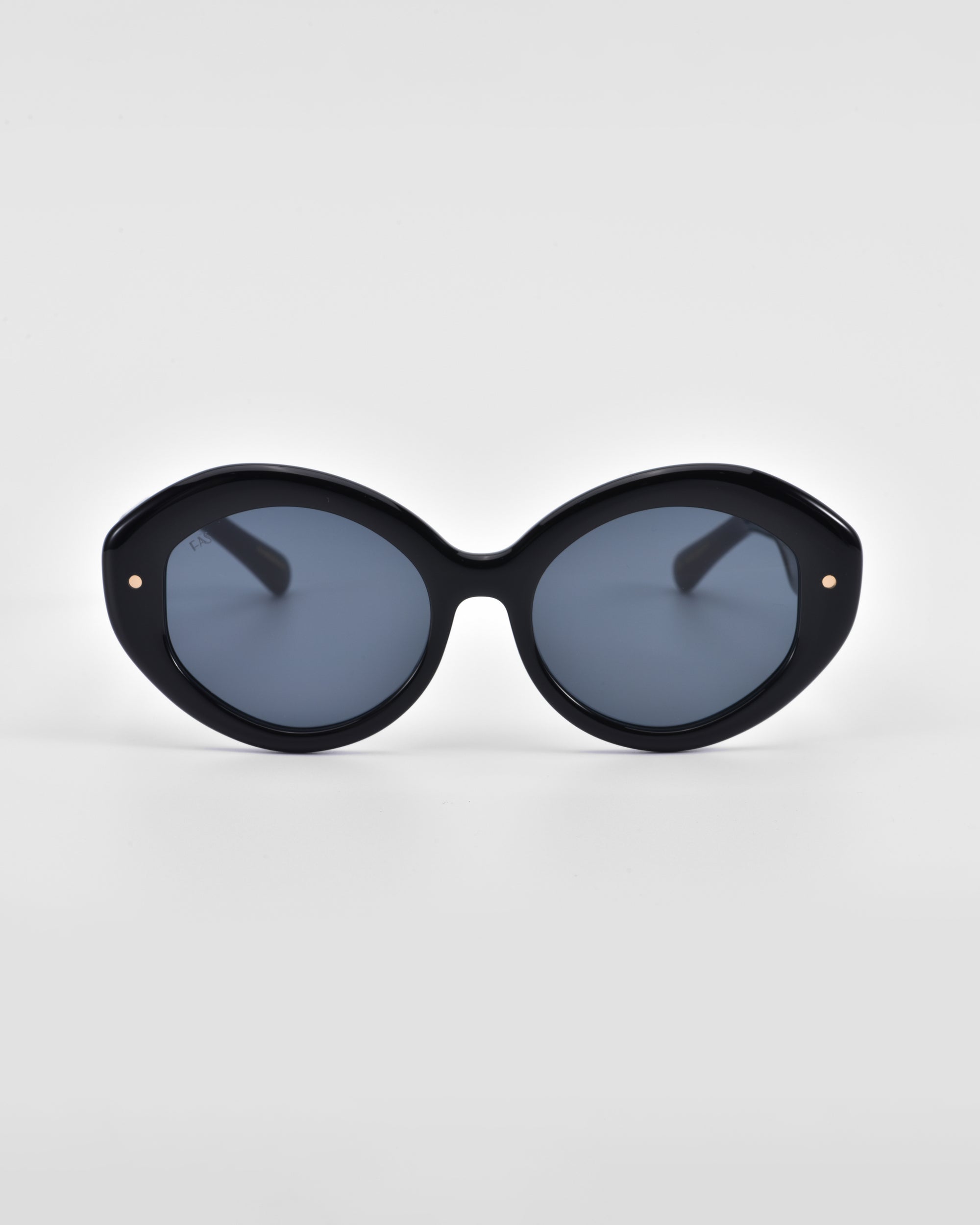 A pair of large, round, black Helios sunglasses with dark lenses by For Art's Sake®. The thick frame features two small metal accents near the hinges, adding a touch of sophistication. This piece of luxury eyewear is set against a plain light gray background.