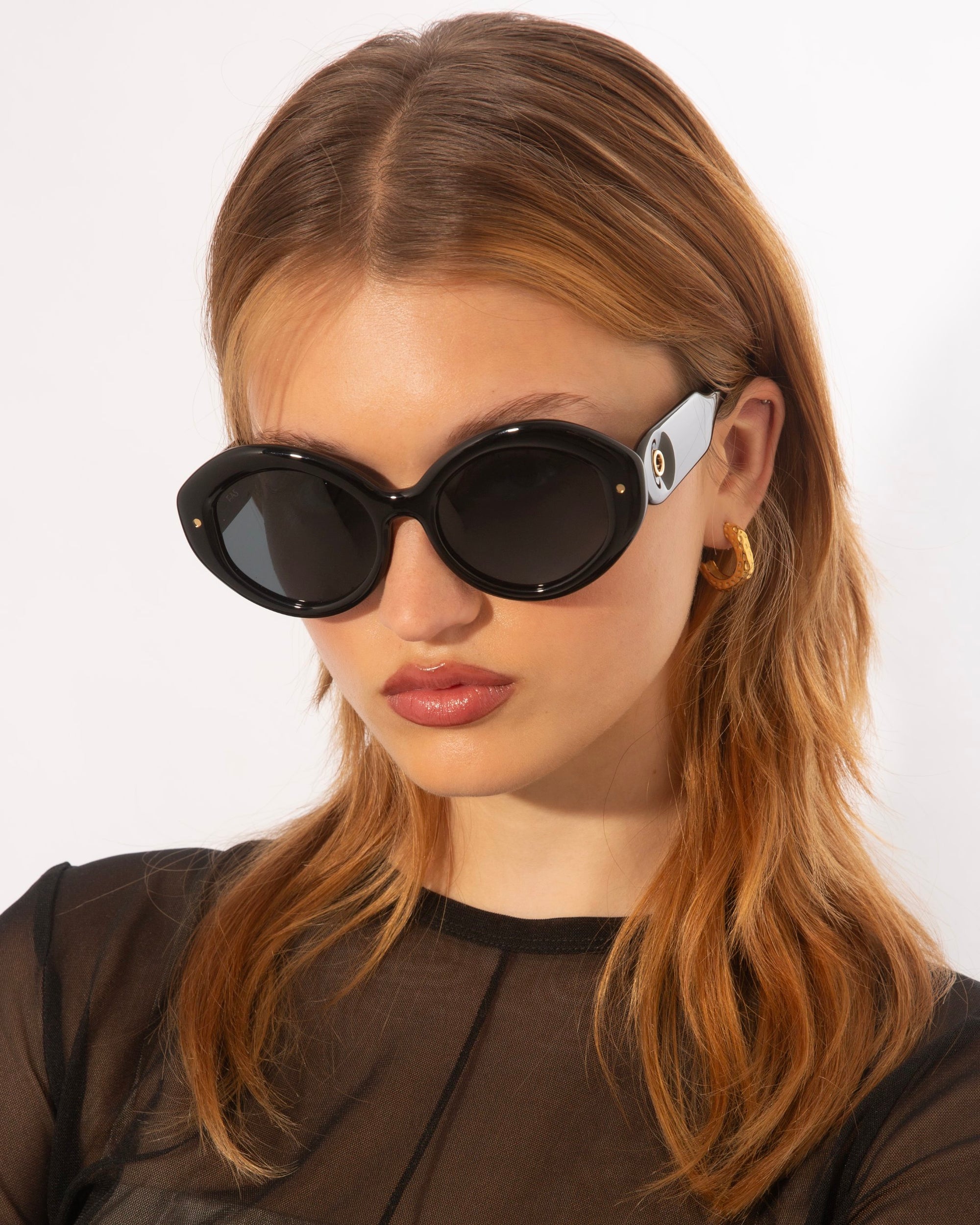 A person with auburn hair is wearing large, black oval-shaped For Art&#39;s Sake® Helios sunglasses that exude luxury eyewear vibes and gold hoop earrings. They have a calm expression and are dressed in a sheer black top. The background is plain white.