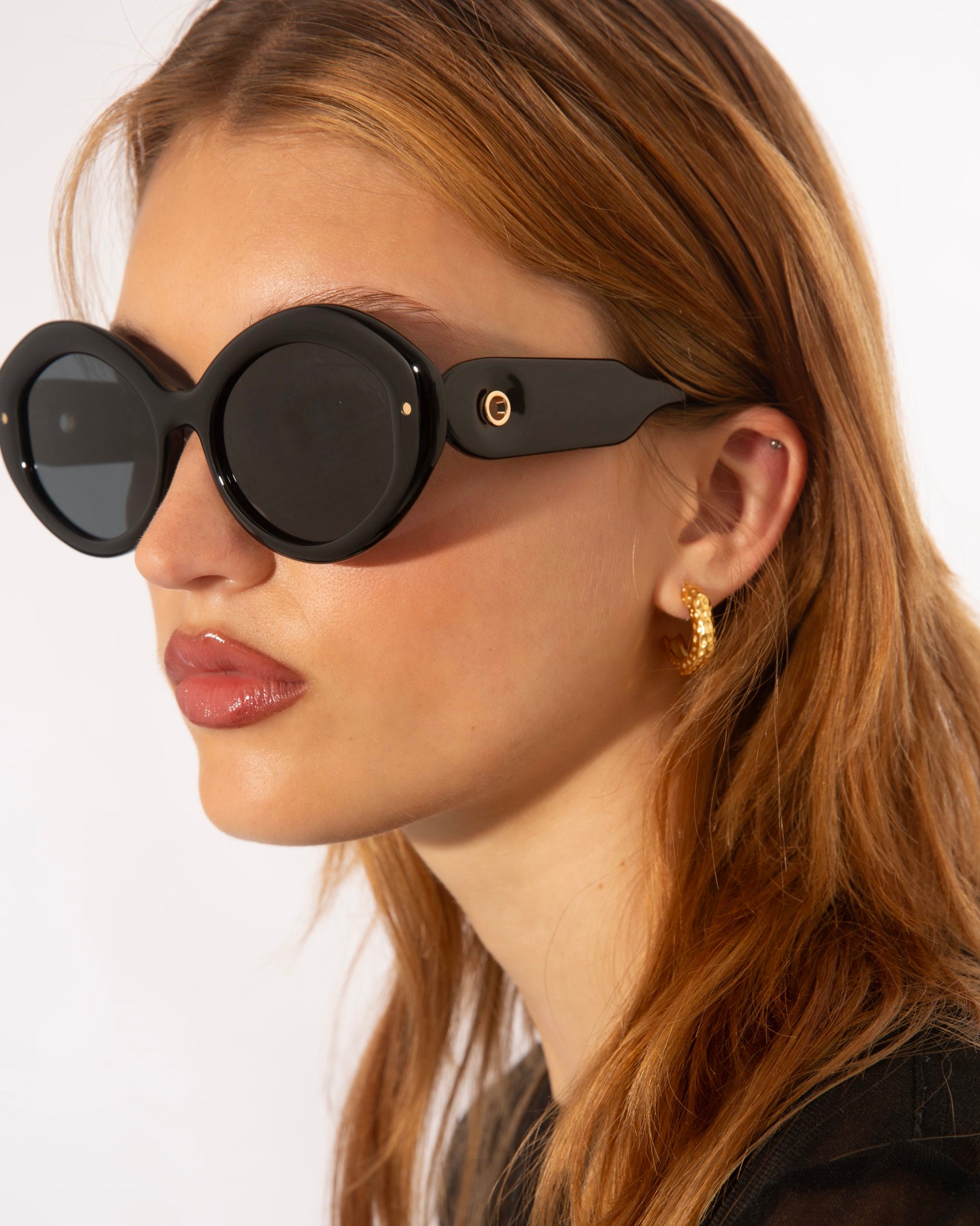 A person with long, light brown hair is wearing large, round black Helios sunglasses from For Art&#39;s Sake® and gold hoop earrings. The background is plain and light-colored, highlighting their luxury eyewear.
