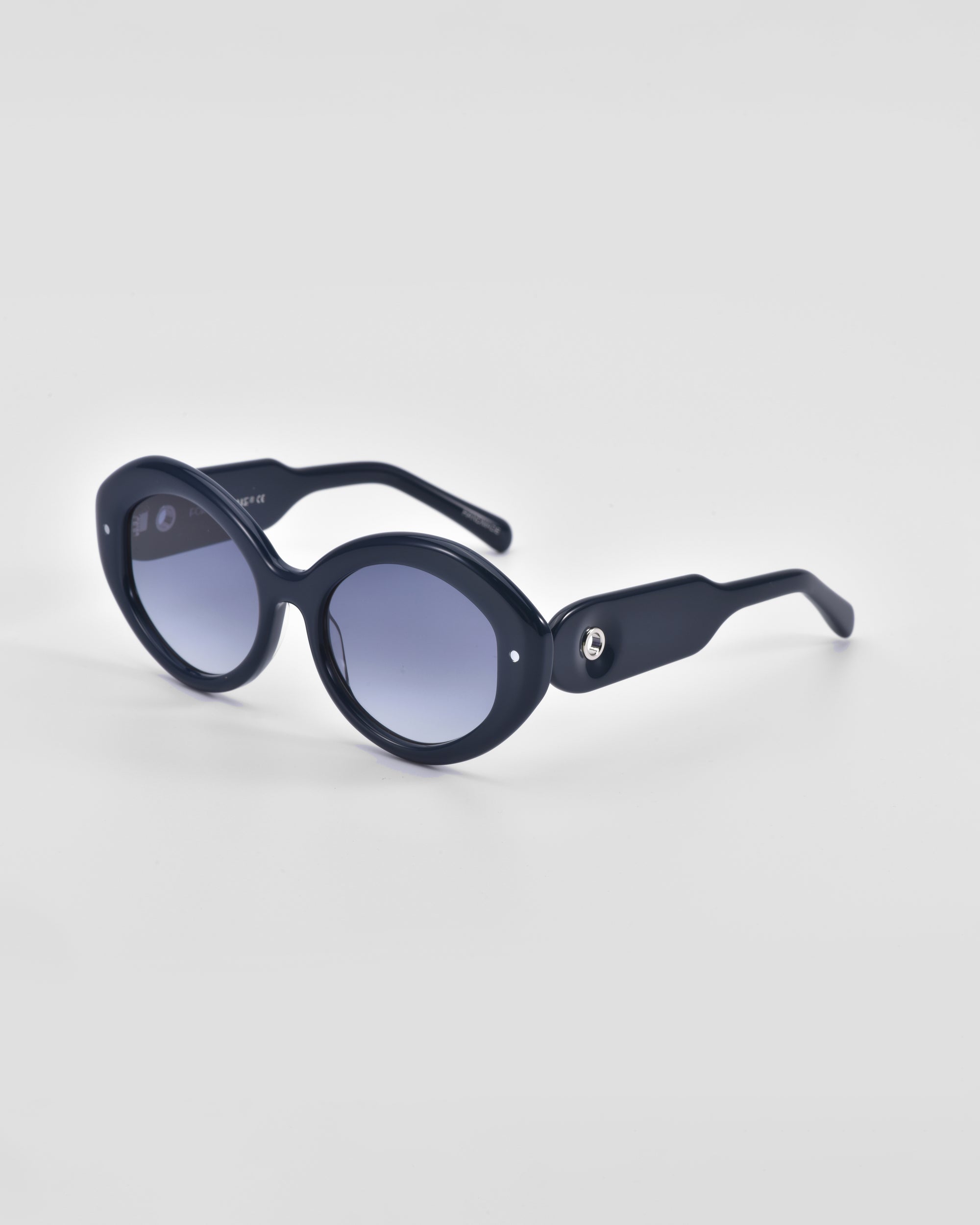 A pair of black oval For Art's Sake® Helios sunglasses with blue-tinted lenses placed on a white surface. The arms are wide and feature a small silver circular detail near the hinge. This piece of luxury eyewear boasts a sleek and modern design, suitable for both casual and formal wear.