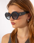 A person with long, light brown hair wears large, round Helios sunglasses by For Art's Sake® that cover part of their eyes and face. They are also wearing drop earrings and a dark sleeveless top, looking slightly to the side against a plain white background.