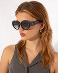 A person with shoulder-length light brown hair is wearing For Art's Sake® Helios sunglasses and a sleeveless gray top. They are looking slightly to the side with a neutral expression. The background is plain white.