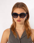 A person with straight, shoulder-length hair is wearing large, oval-shaped Helios sunglasses by For Art's Sake® with a dark blue frame. They are dressed in a sleeveless, grey, v-neck top and have a neutral expression on their face, set against a plain white background.