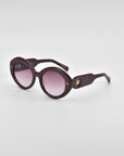 A pair of stylish Helios luxury eyewear by For Art's Sake® with dark, round lenses and thick, glossy brown frames. The arms of the sunglasses are also thick and contoured, featuring small gold embellishments near the hinges. The background is plain white.