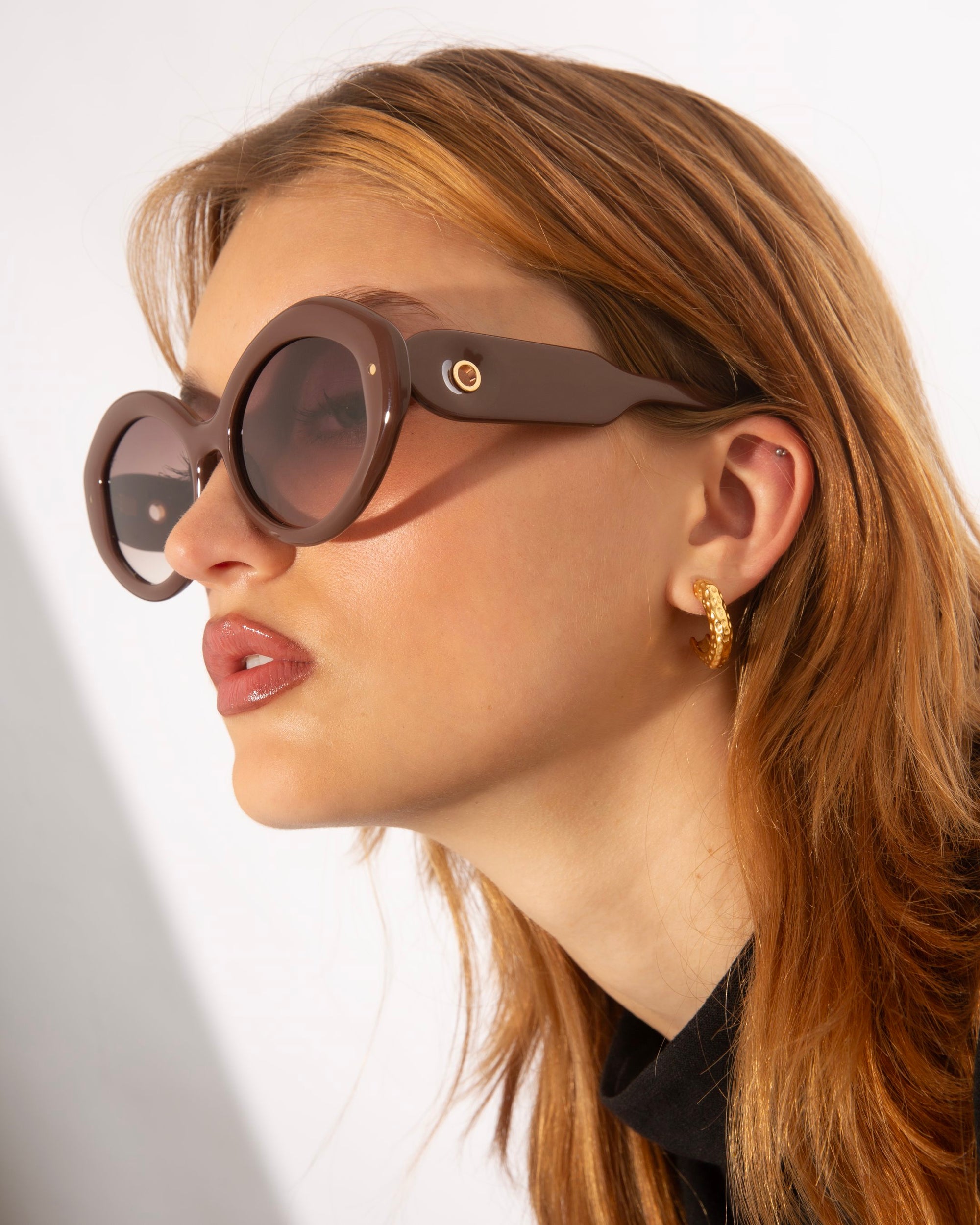 A person with light brown hair wearing luxurious Helios sunglasses from For Art&#39;s Sake® and a golden hoop earring with textured detail. They are dressed in a black top and are looking to the side against a neutral background.