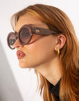 A person with light brown hair wearing luxurious Helios sunglasses from For Art's Sake® and a golden hoop earring with textured detail. They are dressed in a black top and are looking to the side against a neutral background.