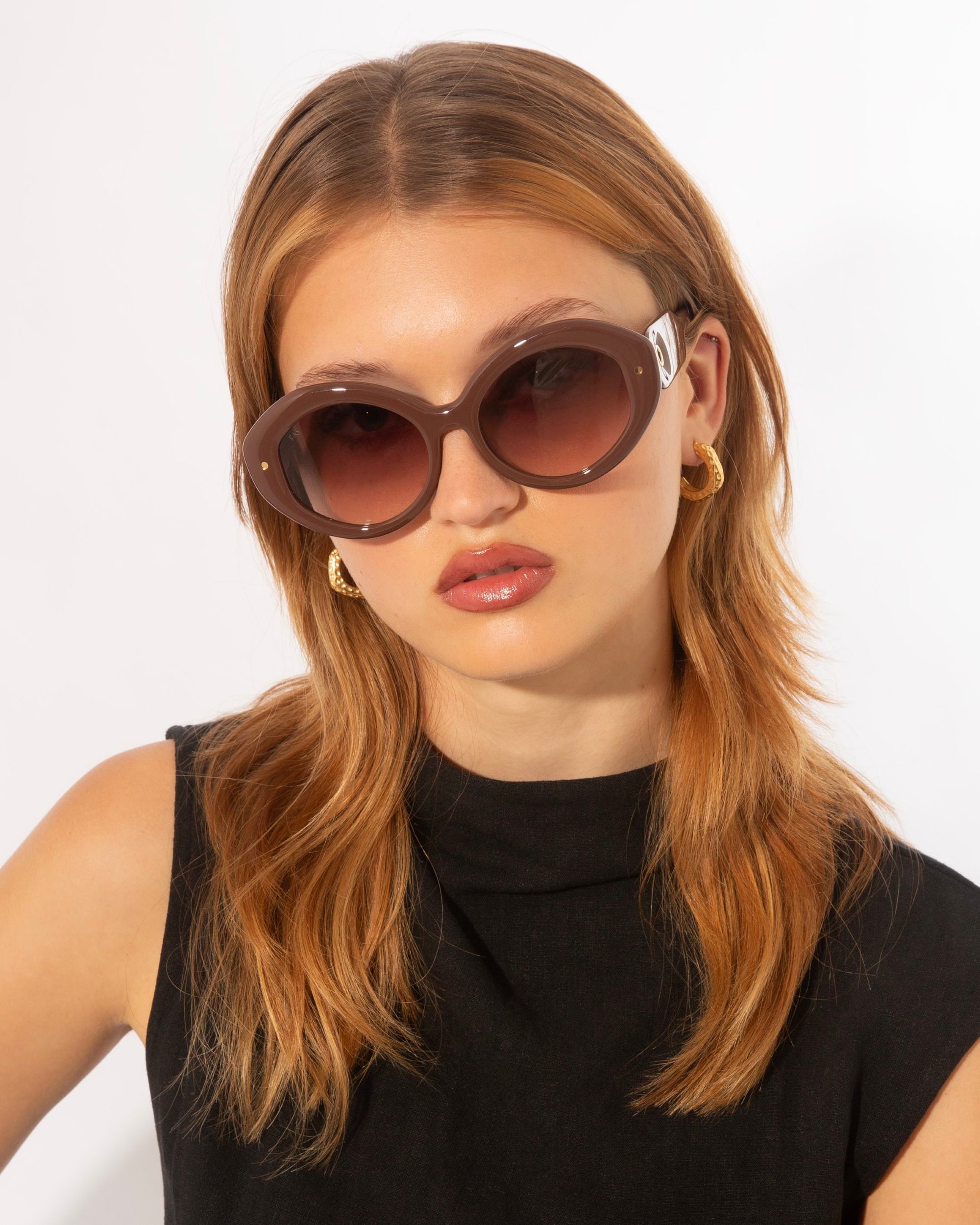 A person with light brown, shoulder-length hair is wearing large, Helios sunglasses by For Art&#39;s Sake® with a brown tint. They have gold hoop earrings and are dressed in a sleeveless black top. The luxury eyewear adds a touch of sophistication against the plain white background.